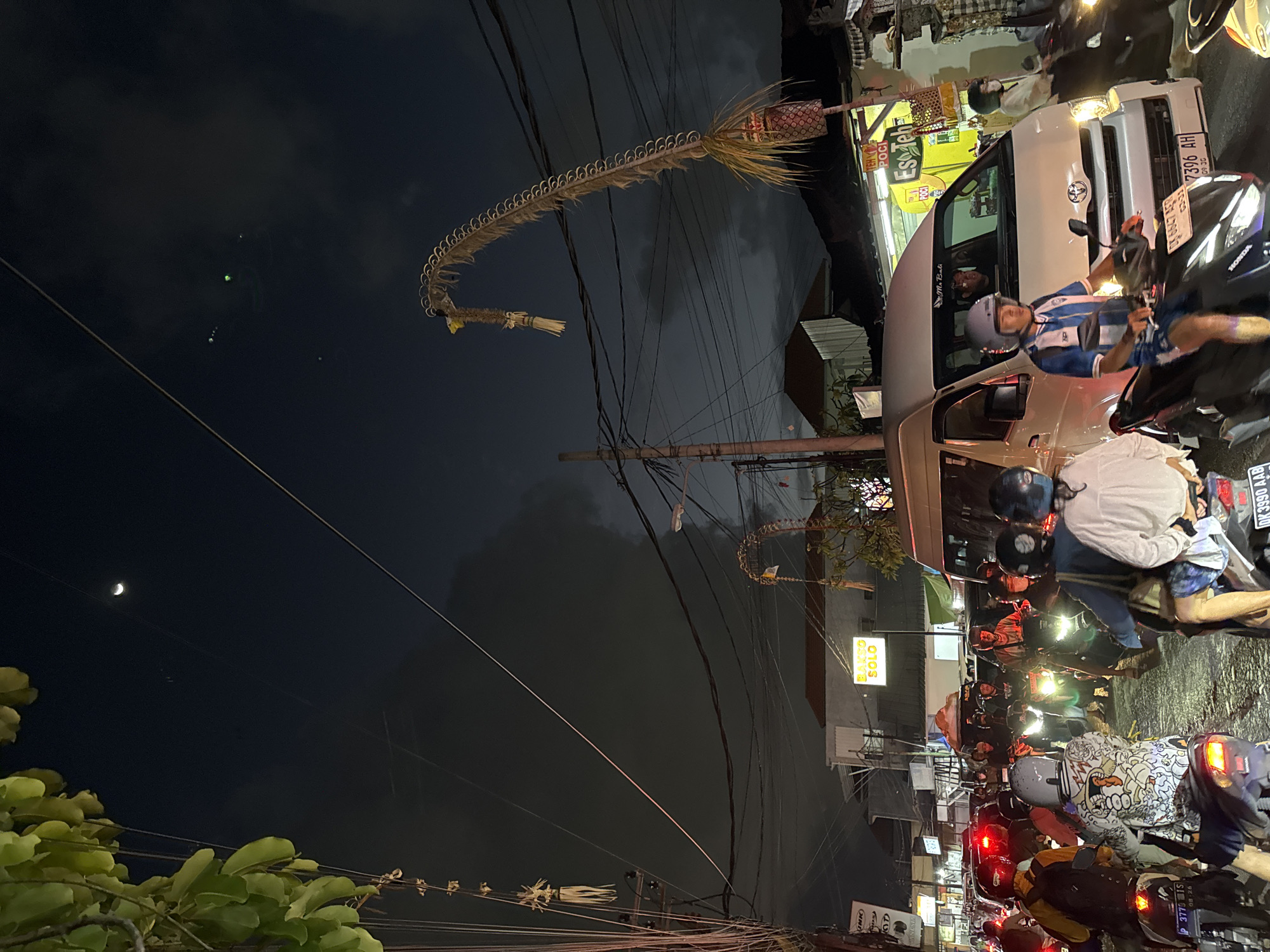 The image depicts a busy street scene at night in Denpasar, Indonesia. A traffic jam is evident, with numerous motorbikes and a white van filling the road. People are riding on the motorbikes, many wearing helmets. The sky is dark with clouds, and a crescent moon is visible. Decorative items, possibly for a festival or cultural event, hang from poles and overhead wires, featuring woven palm leaves. Buildings line the street, with some illuminated signs visible, including one for "BAKSO SOLO". The overall atmosphere is one of urban activity at night, with a strong cultural presence suggested by the decorations.