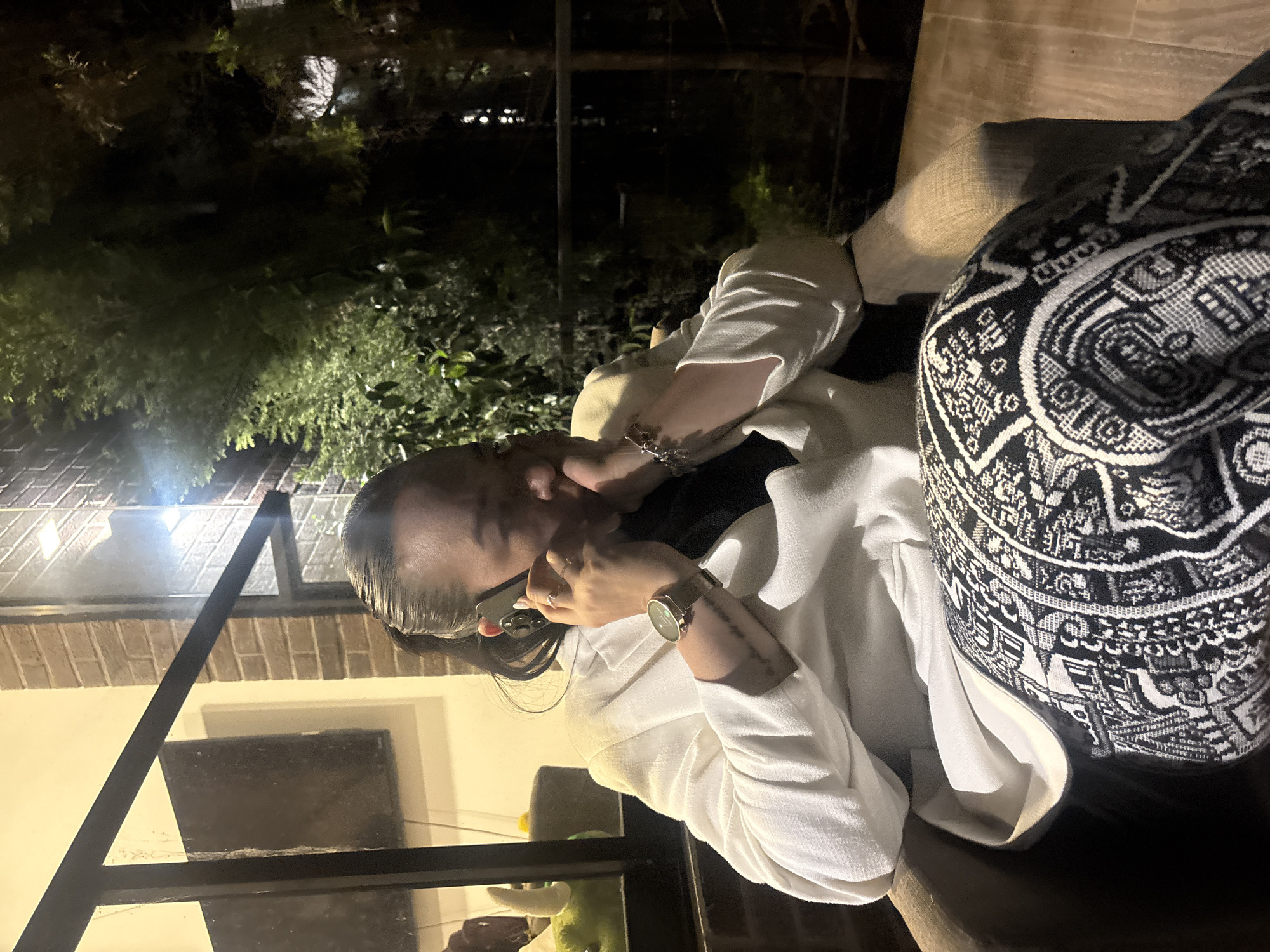 A person is photographed at night, apparently outdoors, in what seems to be San Andrés Cholula, Mexico, due to the general environment and apparent outdoor setting which is a likely indication of restaurants and nightlife common in the city. The individual is holding a smartphone to their ear, possibly on a phone call. They are seated, wearing a white robe, and have a watch on their wrist. A patterned black and white textile object is next to them. Behind them, there is vegetation and structural elements like windows and dark-colored framing. Overall lighting in the scene is dim with contrasting highlights.
