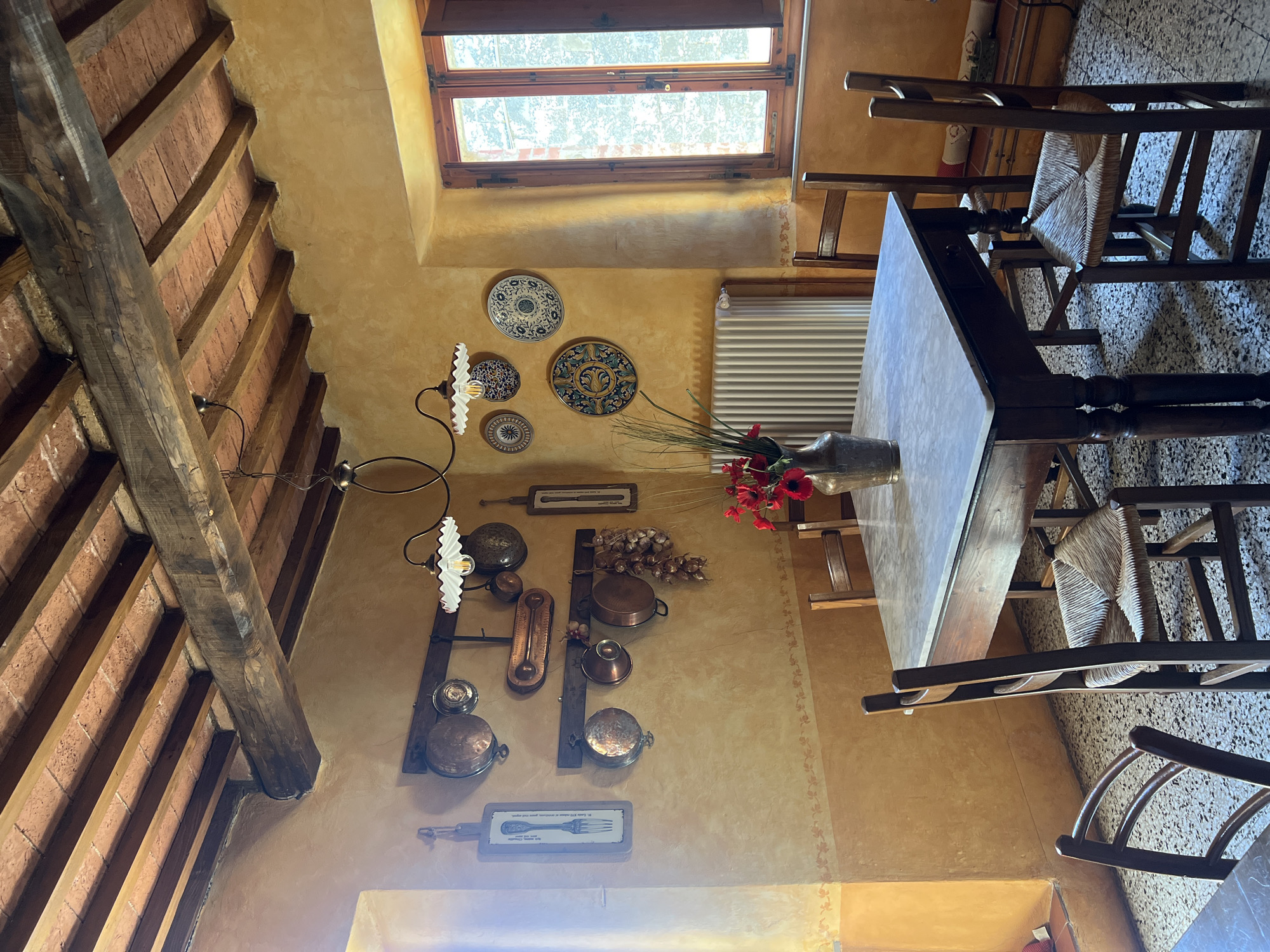 The image captures an interior space in Sinalunga, Italy. The room has a rustic aesthetic, characterized by exposed brick and wooden beams in the ceiling. The walls are painted in a warm, golden hue with decorative floral motifs. Several decorative plates and a collection of copper cookware are mounted on the wall. A table is positioned in the room, and chairs surround it. A vase of red flowers sits on the table. Natural light enters through a window. The floor appears to be tiled.