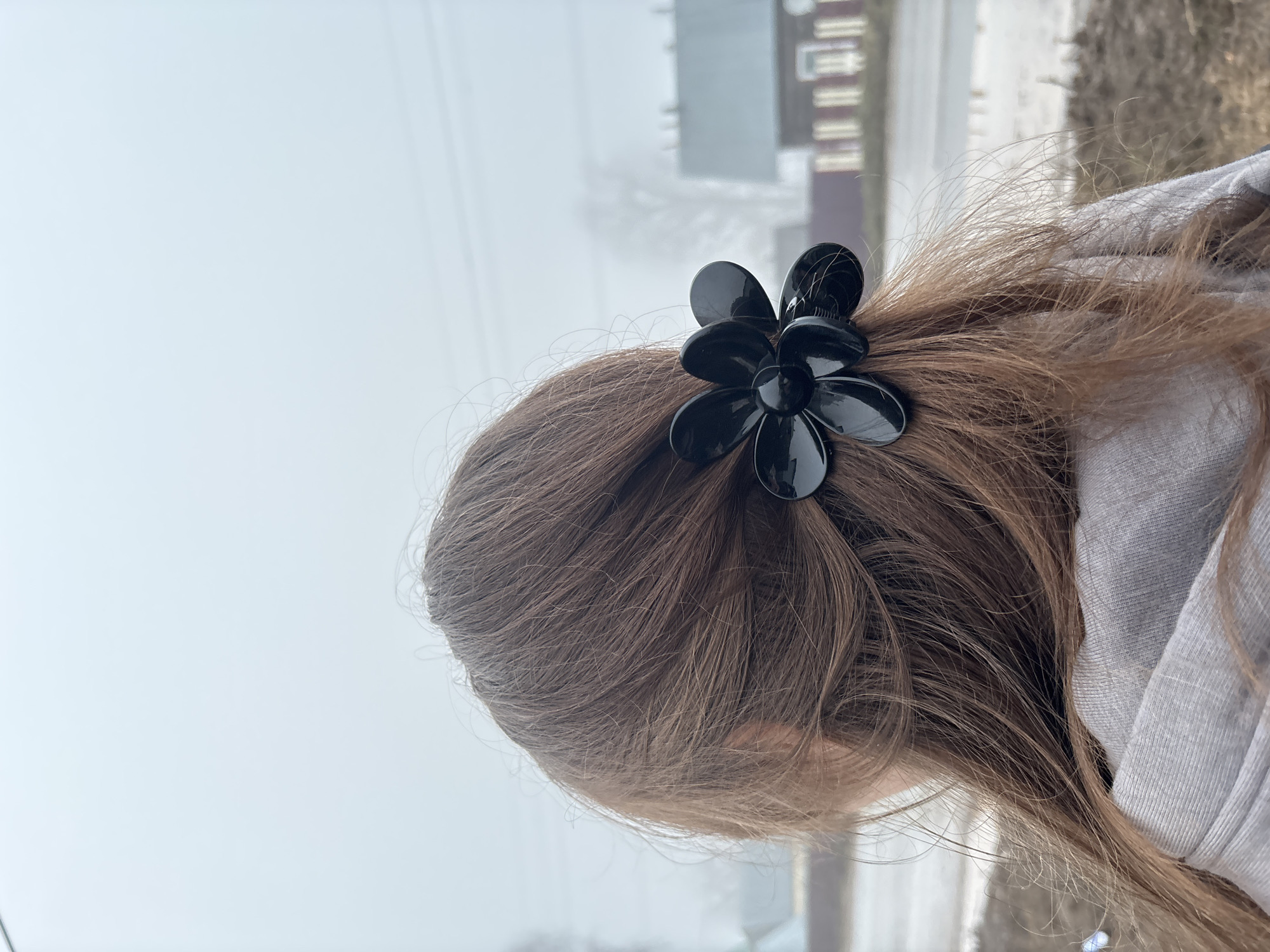 Here's a detailed description of the image:
**People/Subject:** The image features the back of a person's head and hair. The hair is brown, with some lighter strands, and styled with a black flower-shaped hair clip. Part of the person's face is visible, specifically the lower part of their ear.
**Objects:** A black, flower-shaped hair clip is prominent. A gray hooded sweatshirt is also visible.
**Environment:** The background shows an urban environment, with a blurred building in the distance and snow on the ground. The sky is overcast and foggy.
**Location:** The buildings in the distance suggest that the image was most likely taken in Фоки, Russia.