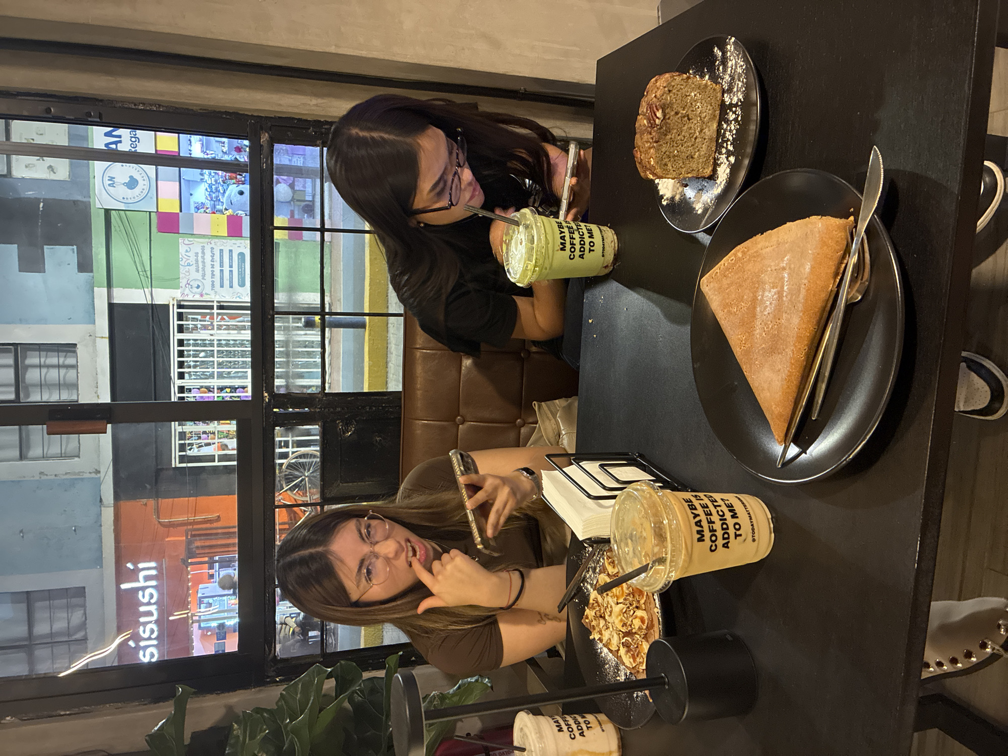 Two young women are seated at a black table indoors. One woman on the left is holding a cell phone with her right hand and pointing to her teeth with her left hand. The other woman on the right is holding a cell phone in her left hand and a straw in her mouth with her right hand. Both women are wearing glasses. The table has food and drinks on it, including a slice of banana nut bread, a slice of cake with silverware, and two plastic cups with the label "MAYBE COFFEE IS ADDICTED TO ME? @TODAYMATTERS". Through the window, a view of the city of San Andrés Cholula, Mexico can be seen. A restaurant with the name "sisushi" is visible through the window. Buildings and signs can be seen in the background.