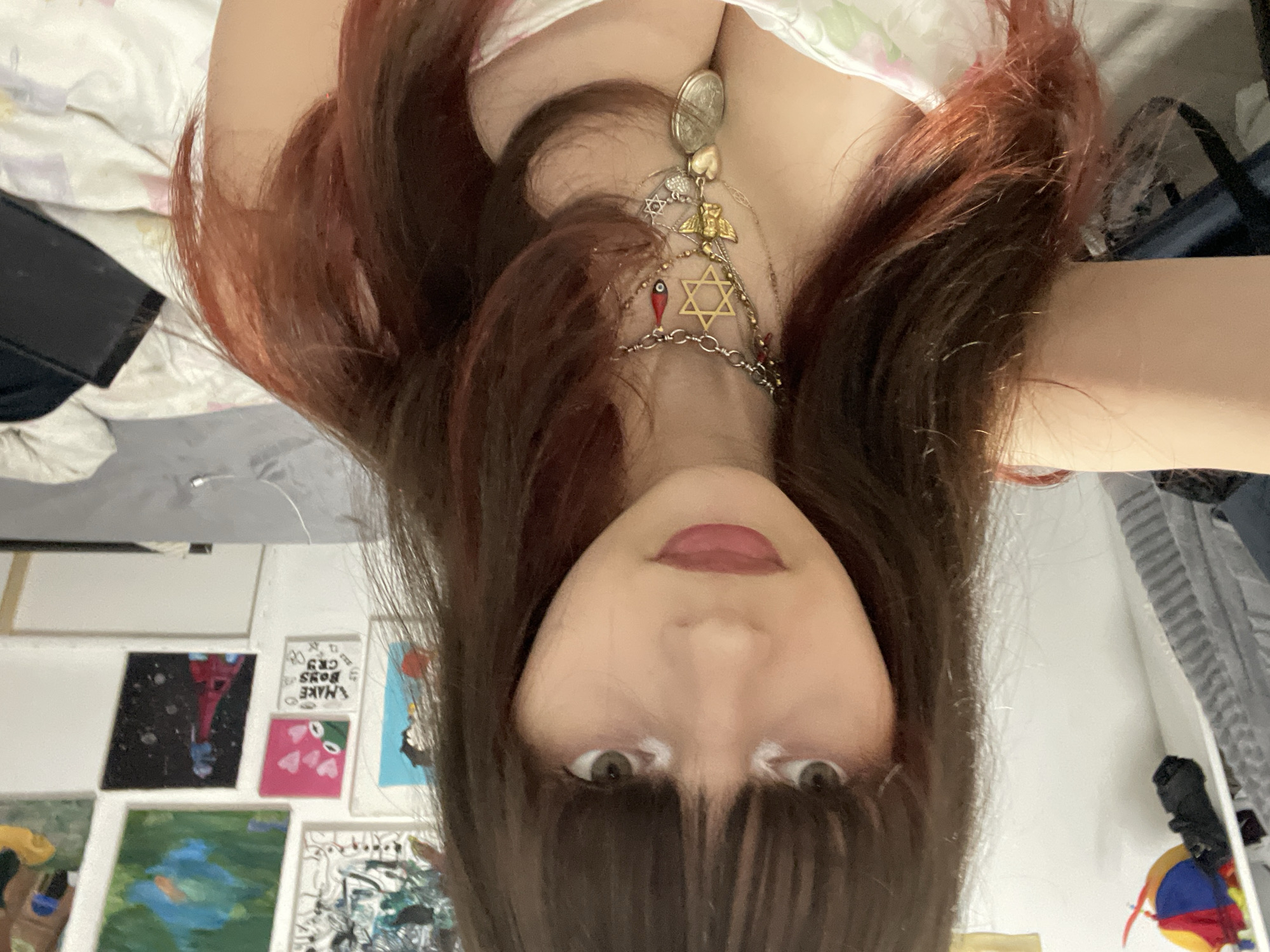 The image shows a person with brown hair and bangs, wearing a necklace with multiple pendants including a Star of David, upside down. The background includes a bed, artwork on the wall, and a closet. There is no information to suggest a specific location, nor does it point to Raanana, Israel.