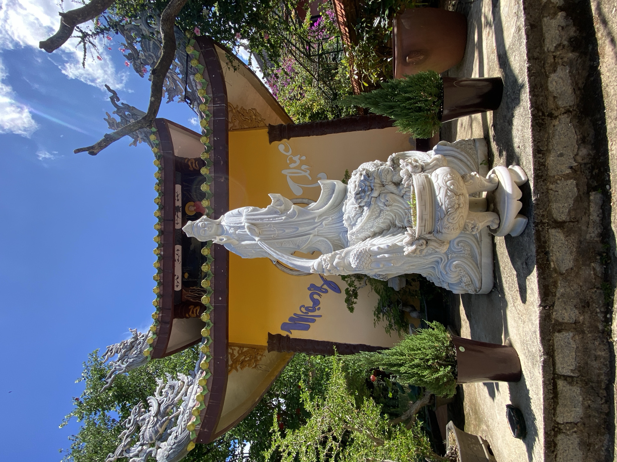This is a full, angled shot of a large, white marble statue of a serene female figure, possibly a deity, with flowing robes and intricate details in the sculpture. She appears to be seated or reclined on a decorative base that resembles water or clouds, with a mythical creature, possibly a dragon or lion, carved into it. The statue is positioned outdoors, in what appears to be a temple courtyard or garden, given the presence of lush green trees and potted plants.
Behind the statue, there is a building with a traditional East Asian architectural style. The building features a yellow facade with blue calligraphy inscribed on it. The roof is adorned with decorative elements, including what look like golden spheres and stylized dragon-like figures.
The scene is bathed in bright sunlight, indicating it is daytime, likely mid-morning or afternoon. The sky is a clear, vibrant blue with a few scattered white clouds. The overall mood is peaceful and spiritual, consistent with a religious or cultural site.
The text in blue on the yellow wall appears to be in Vietnamese. The location is specified as Thôn 5, Vietnam. The presence of traditional architecture, the statue, and the Vietnamese text strongly suggest this is a scene from a Buddhist temple or a similar cultural landmark in Vietnam.