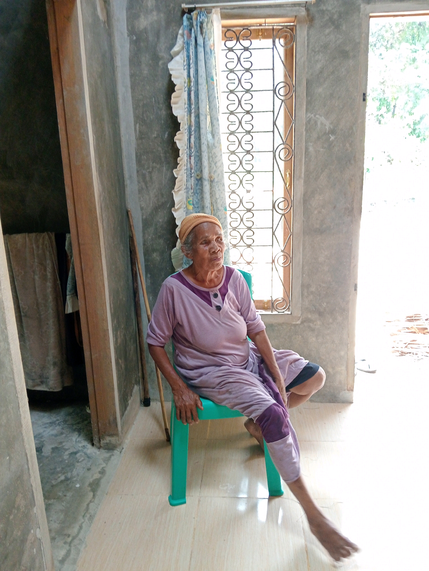 An elderly woman with grey hair and fair skin, wearing a light orange headscarf and a light purple long-sleeved shirt with a darker purple neckline and two dark buttons, is seated on a teal plastic chair in what appears to be a simple dwelling. She is looking off to the right side of the frame. Her legs are crossed, with her left leg extended forward and her right leg bent. Her clothing consists of a light purple garment with darker purple accents on the knee area of her trousers. The environment is dimly lit, with a concrete wall and floor visible. To the left of the woman, a wooden door frame and a dark cloth hanging are apparent. Behind her, a window with a decorative metal grate is present, covered partly by a patterned blue and white curtain. The floor is tiled with light beige and cream-colored squares. The background shows a bright outdoor area with greenery visible through a doorway to the right. A walking stick leans against the wall behind the woman. The image appears to have been taken in Campago Barat, Indonesia, based on the provided context.