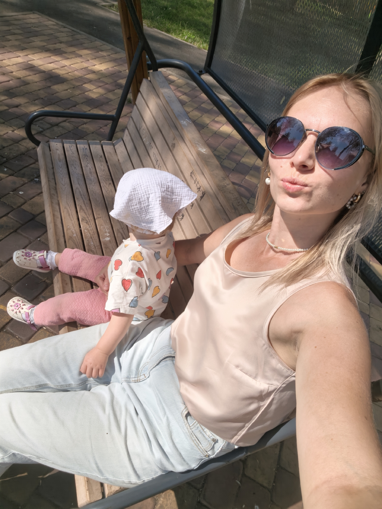 A blonde woman wearing sunglasses, a pearl necklace, and a beige tank top is sitting on a wooden bench swing with a baby. The baby is wearing a white hat, a light-colored shirt with heart patterns, pink pants, and white sandals. The woman is taking a selfie, puckering her lips. The background shows a patterned brick walkway, grass, and a metal railing. The scene appears to be outdoors in a park or public area.