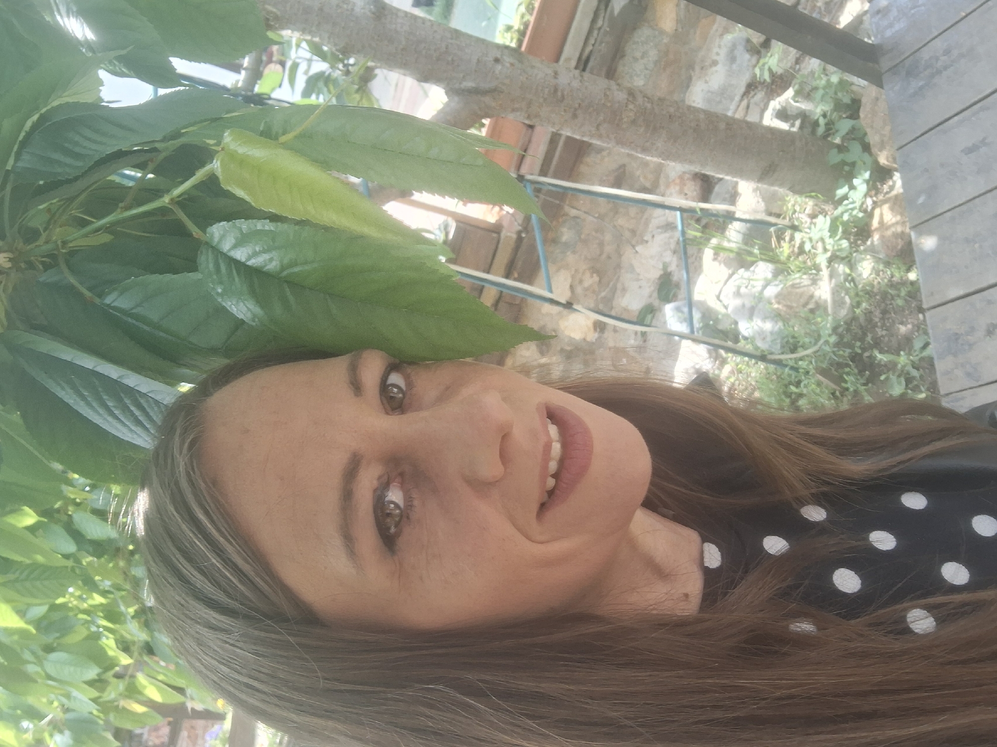 A woman is in Razhdavitsa, Bulgaria. She is smiling and wearing a black and white polka dot shirt. She has long brown hair with gray highlights. She is outside, with green leaves overhead and a wooden deck and stone wall behind her.