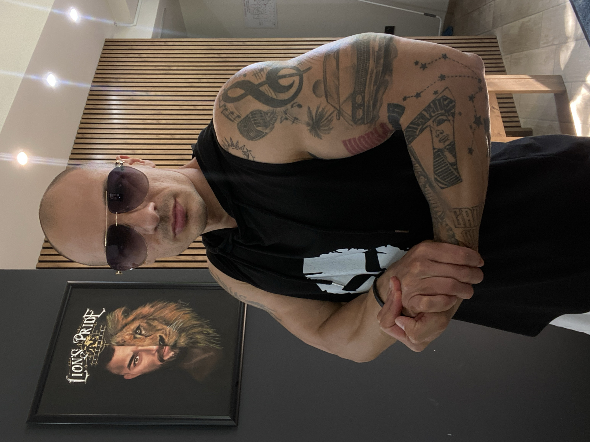 A bald man with sunglasses, wearing a black short-sleeved t-shirt, poses for the camera indoors. The image is oriented sideways. In a corrected view, the man stands upright, looking directly ahead with a neutral expression. He has numerous tattoos visible on both arms; his right arm (left in the image) features a treble clef, an infinity symbol, a microphone, and palm trees, while his left arm (right in the image) displays a pharaoh's head and constellations of stars. A black bracelet is on his left wrist.
The setting appears to be a modern room. Behind the man is a wall with horizontal wooden slat paneling. The ceiling above him is white with several recessed spotlights. To his left, a dark grey wall holds a framed picture. This picture depicts a composite image, half a lion's face and half a man's bearded face, with the text "LION'S PRIDE FITNESS" prominently displayed. To the man's right, a simple wooden chair or bench is partially visible, bathed in natural light suggesting daytime. The ambient lighting combines both natural sunlight and artificial illumination from the spotlights. This scene is located in Pleven, Bulgaria.