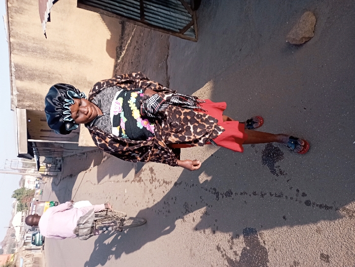 This image captures a daytime street scene in Jos, Nigeria. In the foreground, a dark-skinned woman walks diagonally towards the left, looking downwards. She is wearing a black satin bonnet with a light blue ruffled trim, a grey long-sleeved top, a leopard-print cardigan, a black top with colorful floral patterns visible underneath, a red skirt, and black open-toed sandals with red soles. Her right arm hangs by her side.
In the midground, to the left and behind the woman, a dark-skinned man is riding an older model bicycle uphill, away from the viewer. He wears a light pink long-sleeved shirt and light-colored trousers, with a white package or item secured to the bicycle's rear rack. Both the man and his bicycle cast long, distinct shadows on the asphalt road, indicating strong sunlight from the right.
The environment includes an asphalt road, which appears somewhat uneven and has scattered dark spots or debris. On the far right, a light brown rock rests on the road. Along the right side of the street are single-story buildings constructed with light brown stucco or render, and a section of corrugated metal sheeting. On the left side of the road, further up the incline, more buildings are visible, along with a small green vehicle and a partially visible three-wheeled auto-rickshaw (keke Napep). Distant hills or mountains frame the background under a clear sky.