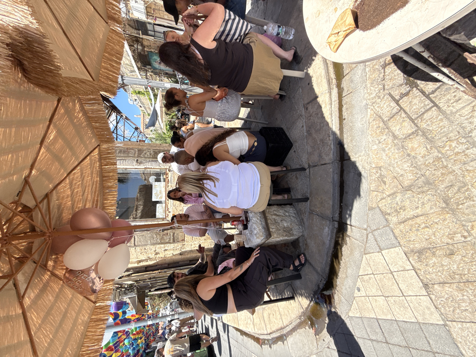 The image depicts an outdoor seating area under a large, fringed umbrella. Several people are seated around a stone table, engaged in conversation and enjoying drinks. The setting appears to be a street or plaza, with a colorful string of decorations hanging in the background, suggesting a festive atmosphere. The architecture and the cobblestone pavement indicate an urban environment. Based on the visual cues and the presence of Hebrew writing on balloons in the cropped image, it is highly likely that this scene is located in Tel Aviv, Israel. The overall impression is one of a casual social gathering in a vibrant city setting.