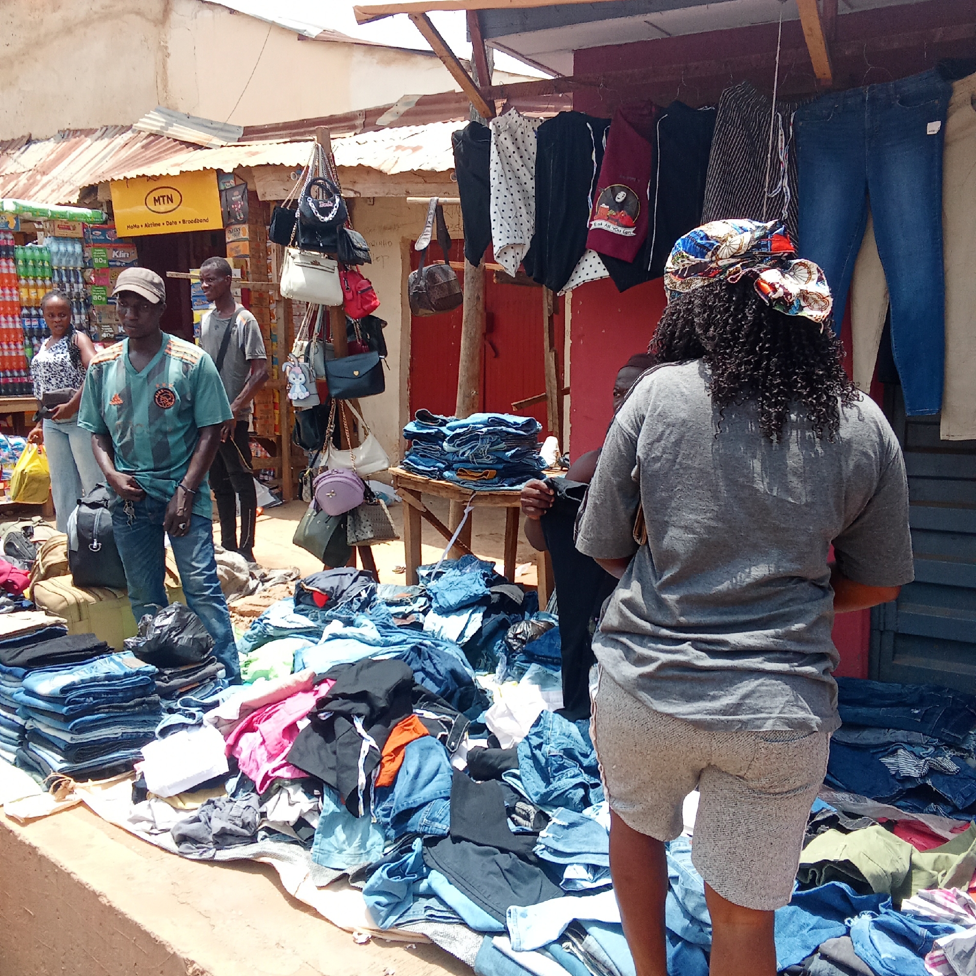 Here is a detailed description of the image:
The image depicts an outdoor market scene in an area with buildings. The location is described as city of Unknown, Unknown. A variety of clothing items, particularly jeans, are displayed for sale on tables and piles on the ground. A man wearing a baseball cap, blue jeans, and a patterned t-shirt is standing near a pile of jeans. A woman wearing a colorful headscarf, gray shorts, and a gray t-shirt is facing away from the camera and appears to be looking at the items for sale. Other individuals can be seen in the background near a store with soda displayed. There are bags hanging on display near the clothing. An "MTN" sign is visible above a store. The area has an open-air market atmosphere with a variety of items displayed.