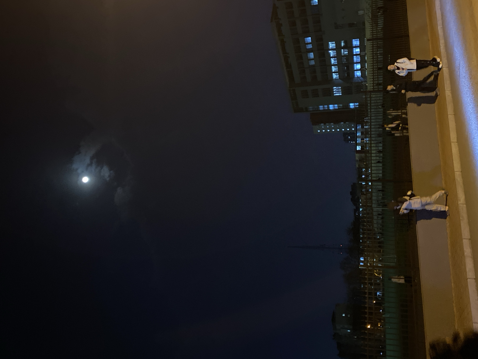 A night scene in Zhongtun, China, featuring a clear sky with a visible moon. Several individuals stand on the side of a road next to a tall building. The building has multiple lit windows. A fence runs parallel to the road. Distant trees and buildings are visible in the background.