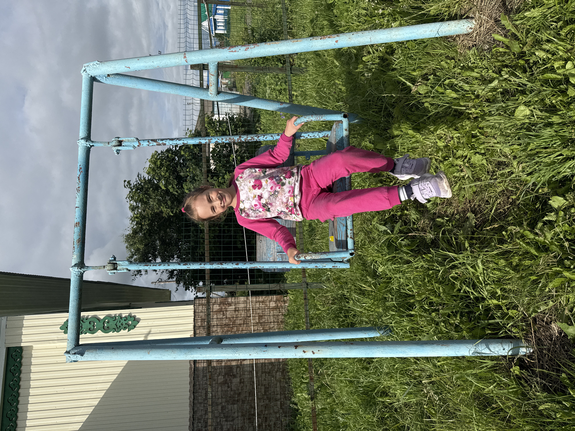 A young girl with brown hair, wearing a pink floral shirt and pink pants, lies on a blue metal structure in an outdoor setting. She is lying on her back, with her right arm bent and her hand resting on the metal. Her eyes are closed, and she appears to be relaxed or sleeping. The metal structure looks like a swing set or a part of a playground, painted light blue with some rust showing through.
The girl is positioned on a horizontal bar of the structure, with her legs extended. She is wearing grey sneakers with white laces and some black and white striped socks. The background consists of green grass and trees, suggesting a park or a rural area. The sky is overcast with grey clouds, indicating a cloudy day.
In the background, there are glimpses of buildings, possibly a house or sheds, with white and light yellow siding. There is also a metal fence and more green foliage. The overall scene is casual and relaxed, with the girl seemingly enjoying a moment of rest outdoors. The time of day is difficult to ascertain precisely due to the overcast sky, but it appears to be daytime. No visible text is present in the image. The location is specified as Bolshoy Turysh, Russia.