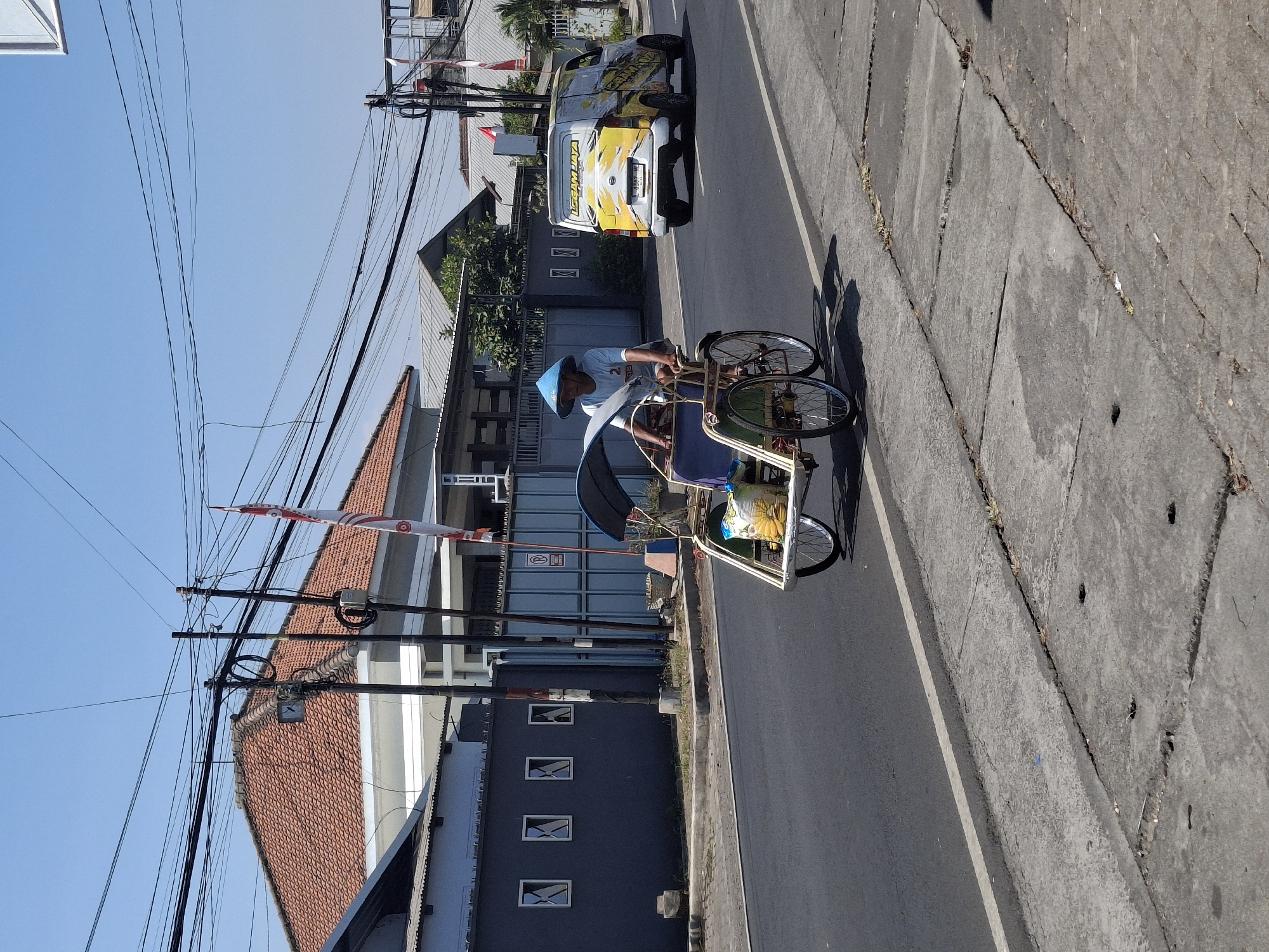 In Lumajang, Indonesia, a man wearing a conical blue hat pilots a three-wheeled pedicab along a paved road under a clear blue sky. The pedicab is a traditional mode of transport, with a covered passenger area and a cargo area at the back. The man is seated and pedaling, facing forward. The pedicab contains a yellow bag with a sunflower design and other items.
To the right of the pedicab, a white and yellow van with graphics featuring a sunburst and text travels in the same direction. The van has a license plate that reads "L 9387 HC."
The background reveals residential and commercial buildings. On the left, a house with a reddish-tiled roof and white walls is visible, partially obscured by numerous overhead power lines. A dark gray wall with three framed rectangular windows is also present. On the right, more buildings with roofs and trees can be seen. The street is lined with a concrete sidewalk. The overall scene depicts a typical day in a busy Indonesian city, with local transportation and modern vehicles sharing the road.