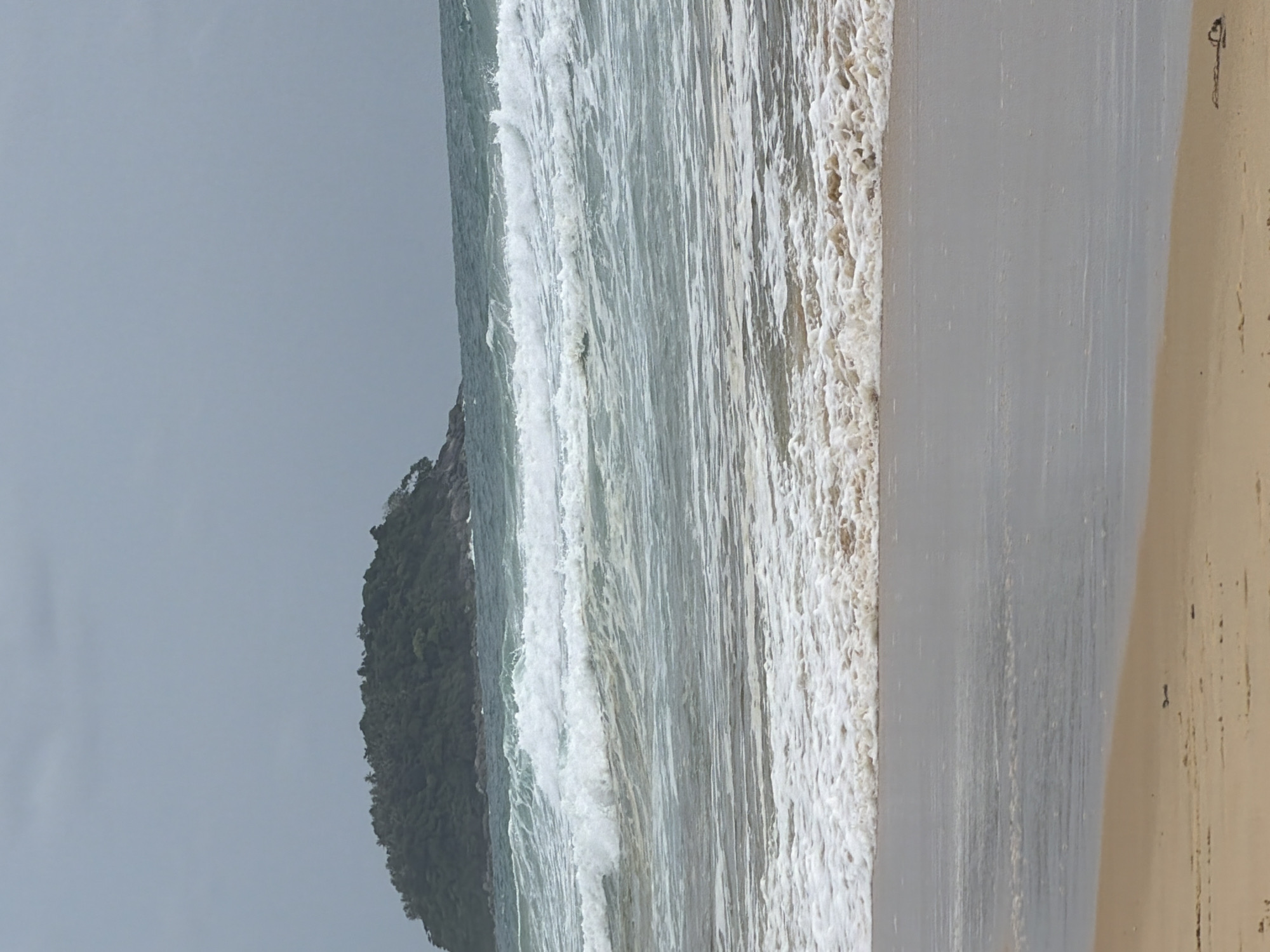 The image depicts a tropical beach scene, likely in Karon, Thailand, on an overcast day. In the distance, a lush, tree-covered island rises from the turquoise ocean. The foreground features a sandy beach, with waves crashing onto the shore, creating a foamy white surf. The water is a mix of blue and green hues, with choppy waves indicating a moderate surf. The sky is a uniform gray, suggesting cloudy weather. No people or man-made structures are visible in the frame, emphasizing the natural beauty of the coastal landscape.