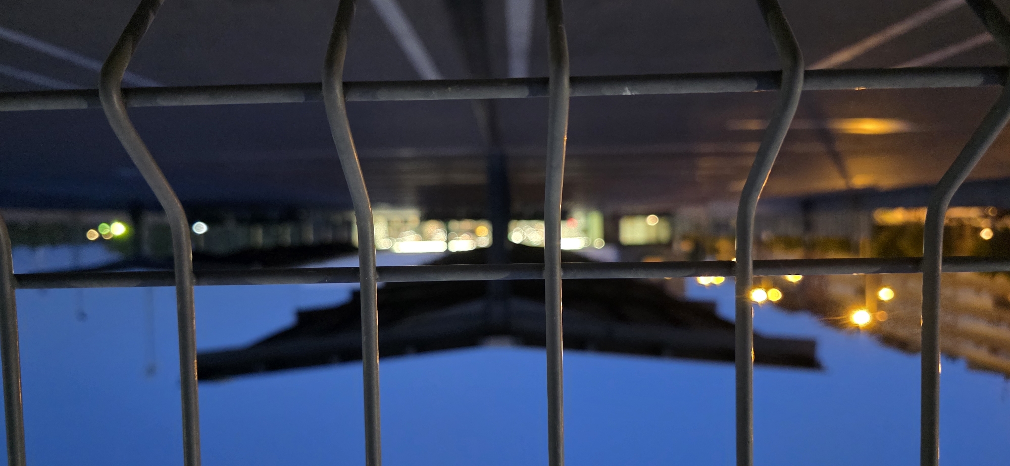 The image depicts a wire fence in the foreground, with its metallic bars obscuring the background. Beyond the fence, at dusk or early night, is a blurred scene of an open area, possibly a parking lot or a public space. Several streetlights with a warm, yellow-orange glow are visible on the left side of the image, illuminating a building facade and casting reflections on the ground. In the middle ground, a large, dark, roof-like structure with an unusual, wing-like silhouette is prominent. Beneath this structure, there appear to be illuminated storefronts or buildings, with lights creating soft, bokeh effects. White lines, indicative of parking stalls or road markings, are faintly visible on the dark ground. The sky is a deep blue, transitioning to a darker hue at the top. The overall atmosphere is dim and somewhat indistinct due to the low light conditions and shallow depth of field. The specific location within Huesca, Spain, is not discernible from the visual elements present in the image.
