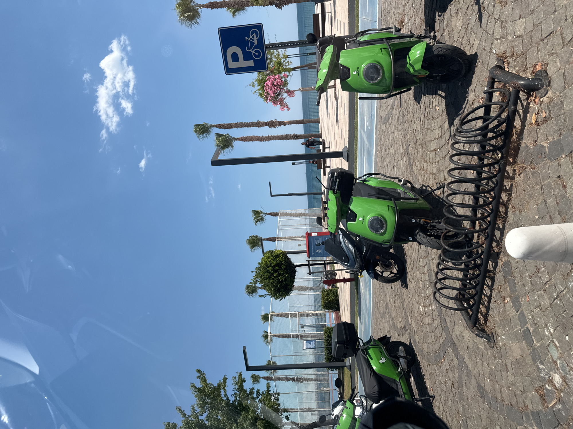 The image shows a bicycle parking area in Konyaaltı, Turkey, near the beach. There are three green Marti-branded scooters parked near a metal bicycle rack. Behind the scooters, there is a walkway with palm trees, a blue and white bicycle parking sign, and the sea. The sky is blue with a few white clouds. The ground is paved with gray stones.