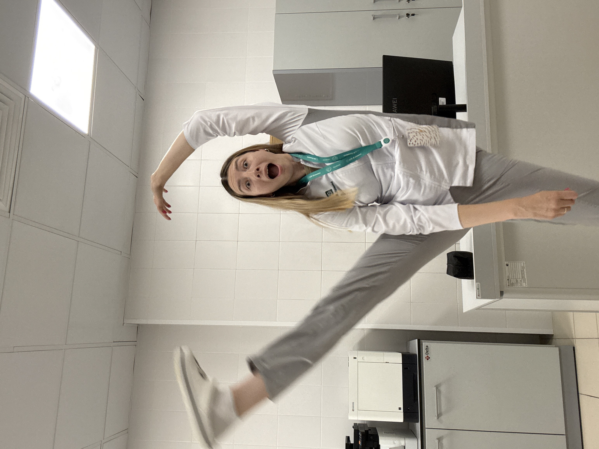 A young woman with blonde hair is captured in a dynamic pose, with her right leg extended high into the air. She is wearing a white lab coat over grey trousers and white sneakers. Her left arm is raised above her head, and her mouth is open in a surprised or excited expression. She appears to be indoors in what looks like a laboratory or office setting. The walls are tiled in white, and the ceiling has fluorescent lighting panels. In the background, there are laboratory equipment, cabinets, and a computer monitor. The overall impression is one of playful energy within a professional environment. The specific location in Moscow, Russia is not determinable from the provided image.