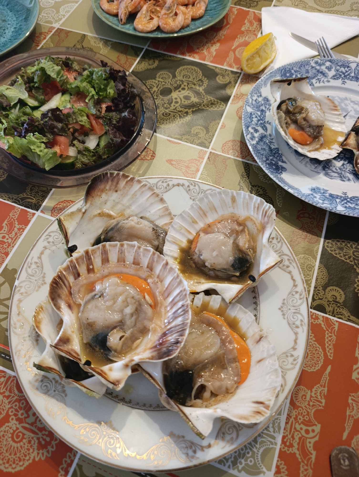 An overhead shot shows a meal laid out on a tiled table. In the foreground, a white plate with a gold rim holds three large scallops in their shells, cooked with some liquid. Behind this, another white plate with a blue floral pattern holds a single scallop. To the left, a glass bowl contains a salad with lettuce, cucumber, and tomato. Further back, a green plate holds several cooked shrimp. A slice of lemon rests on the table near the blue and white plate, and a fork and knife are visible on a white napkin in the upper right corner. The table itself has a patterned surface. The scene appears to be from a domestic setting, likely a home in Setúbal, Portugal, given the presence of seafood and the overall presentation. There are no people visible in the image.