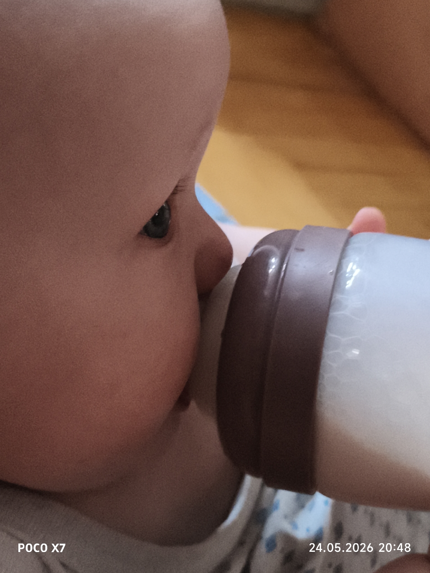 A close-up shot depicts a baby drinking from a bottle. The baby's face is in profile, showing a chubby cheek, nose, and one eye. The bottle is held in the hand of a person, and its nipple is in the baby's mouth. The bottle contains a white liquid. The background is blurry, but appears to be a room with wooden elements. At the bottom of the image, "POCO X7" and "24.05.2026 20:48" are printed, likely indicating the camera and date/time the picture was taken. Location: Unknown, Unknown.