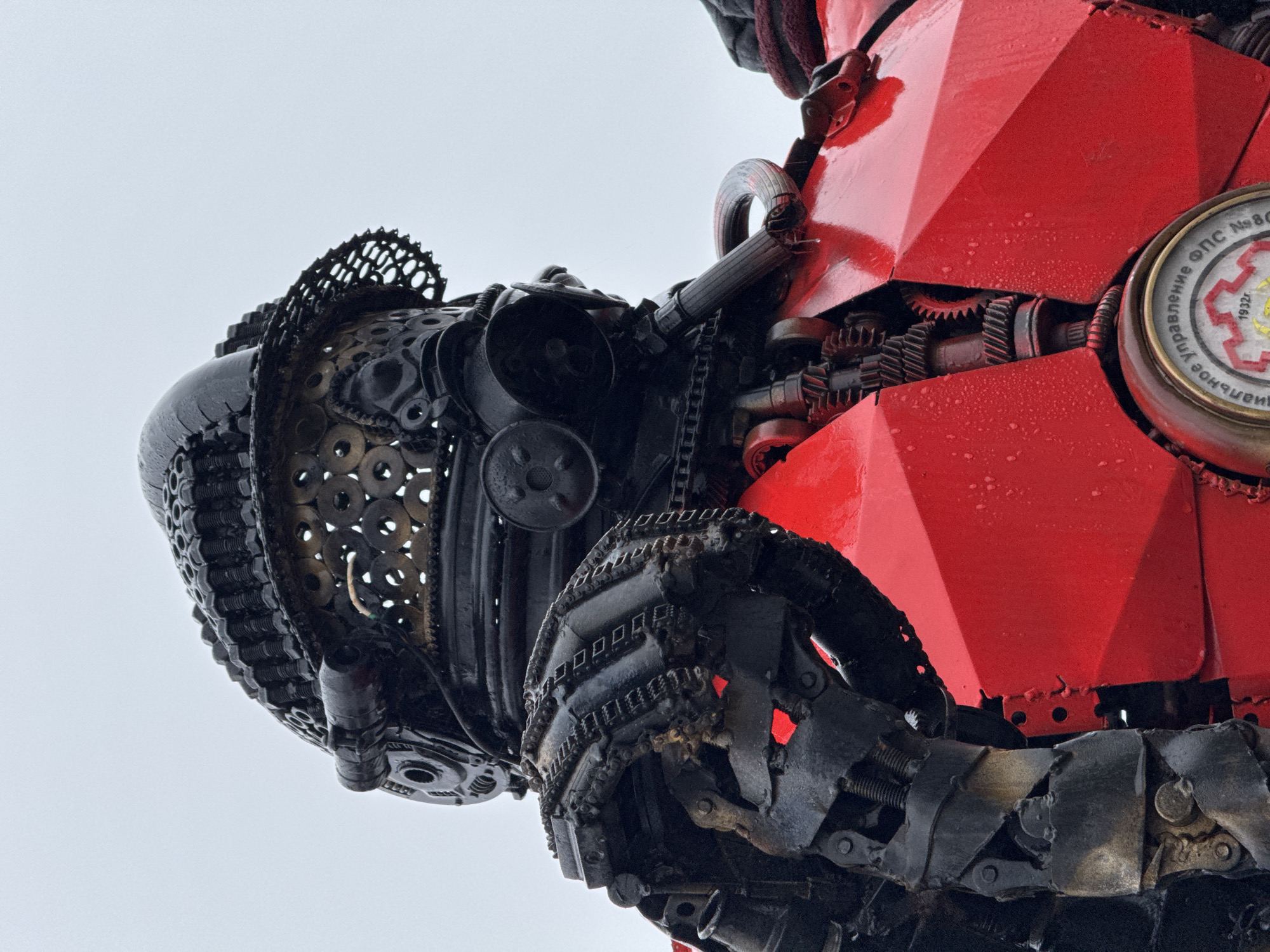 The image shows a close-up of a large metal sculpture located in Votkinsk, Russia. The sculpture appears to be of a humanoid figure, constructed from various pieces of scrap metal, including gears, chains, and other mechanical components. The head of the sculpture is rounded and covered in washers and other metallic circular pieces, and a row of chains with metallic rectangular pieces around the bottom of the head. The body includes sections painted bright red, which appear to be made of sheet metal welded together. A round, clock-like device is attached to the side of the red body panels, and a network of gears and rods is visible behind the red plating. The background is a light gray overcast sky. Overall, the sculpture has an industrial, steampunk aesthetic.