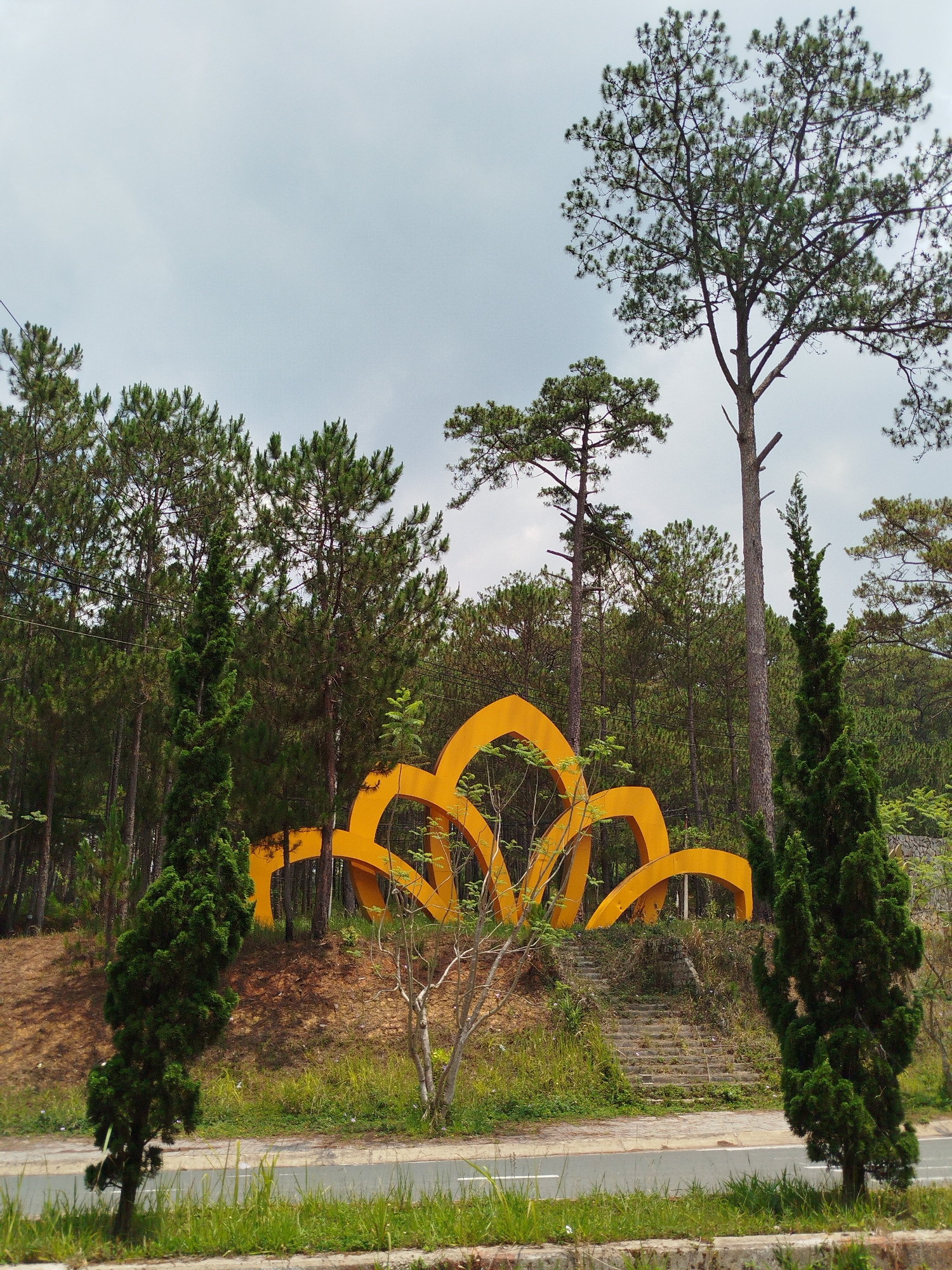 A roadside view in Da Lat, Vietnam presents a striking yellow art installation resembling a stylized lotus flower nestled on a grassy slope, set against a backdrop of dense pine trees under a cloudy sky. A small bare tree and two tall, thin evergreen trees flank the art piece near the road. Concrete stairs lead up the slope towards the sculpture. The roadside is demarcated by a grassy verge and a concrete curb. Power lines are visible in the background.