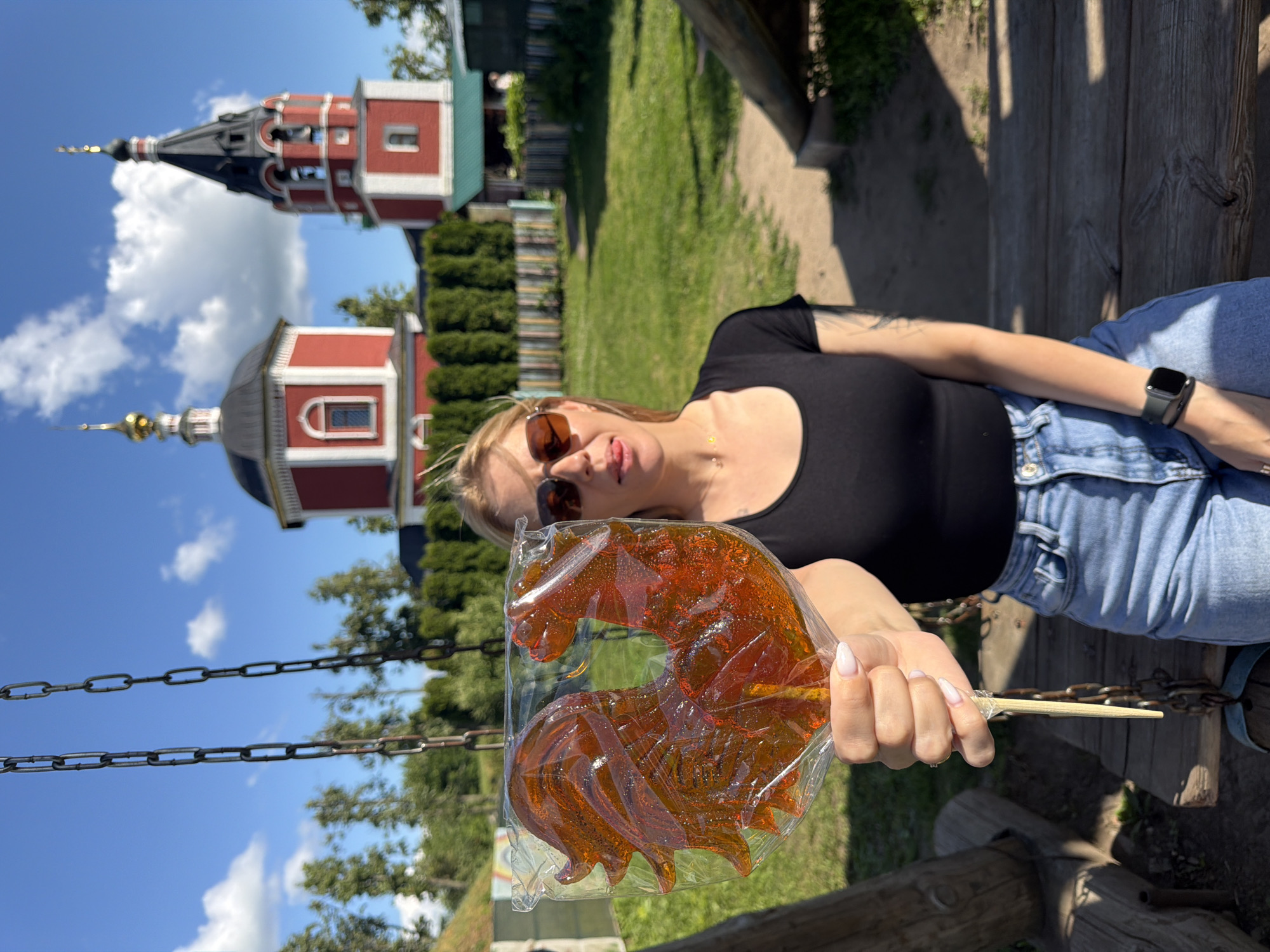 A woman is sitting on a wooden swing in Suzdal, Russia, holding a large, rooster-shaped lollipop. The lollipop is made of translucent amber-colored candy and is wrapped in plastic. The woman is wearing a black t-shirt, blue jeans, and sunglasses. She is sticking her tongue out playfully. In the background, there are two red and white Russian Orthodox churches with golden domes and a black spire. The churches are surrounded by green trees and a wooden fence. The sky is blue with some white clouds. The scene is set in a park or historical area.