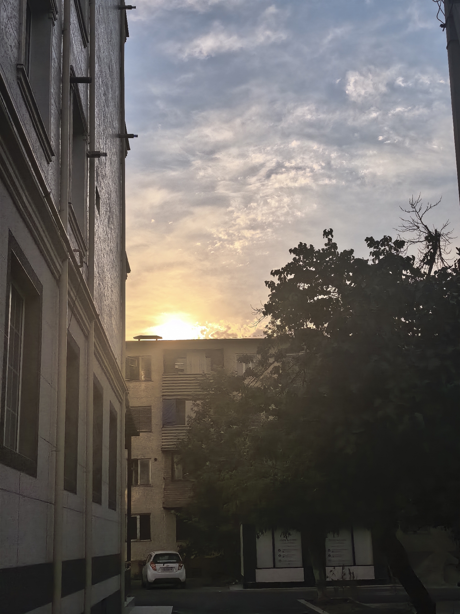 A low-angle, eye-level shot depicts a street scene in Fergana City, Uzbekistan, at dusk. The image is dominated by the left side of a light-colored, textured building with multiple windows and decorative moldings, casting a shadow. To the right, a silhouetted tree with dark leaves and bare branches stands in front of a multi-story apartment building. The sun is low on the horizon, peeking through clouds that are illuminated with warm orange and yellow hues, suggesting an approaching sunset. The sky above is a mix of blue and grey with scattered white clouds. In the foreground, a white car is parked on the street, and two signs are visible behind the tree. The overall impression is one of an urban environment transitioning into evening.