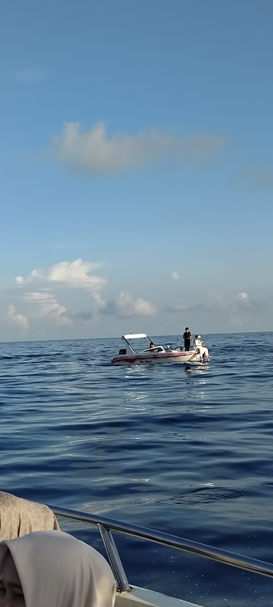 In the foreground, the edge of a boat is visible, with a person wearing a light-colored headscarf and a beige garment. The boat's metal railing is prominent. In the middle ground, a small, white and red motorboat is on the sea. Three individuals are on this boat. One person is standing in the middle of the boat, and another is seated at the back, facing away from the viewer. A third person appears to be seated in the front of the boat. The sea is a deep blue with noticeable ripples. In the background, the sky is a clear light blue with scattered white clouds, suggesting a bright, possibly daytime scene. The overall environment is oceanic, with a sense of calm water and a clear sky. This scene likely depicts a leisurely activity or transportation on the water in or near Sabang, Indonesia.