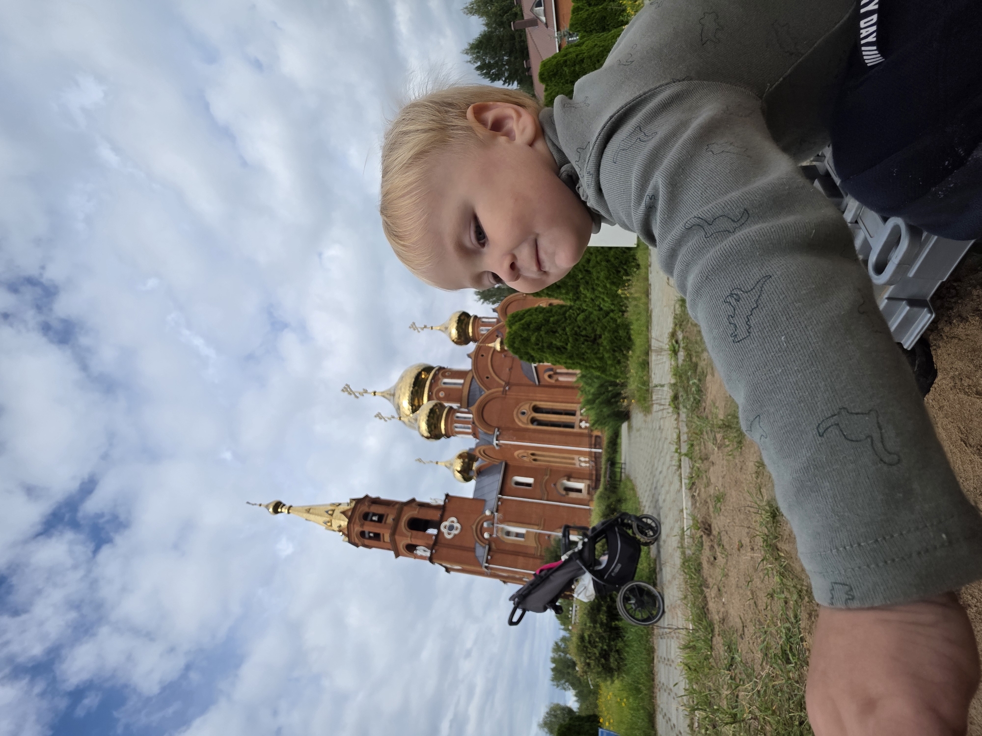 In an outdoor, ground-level shot, a young blonde child in a gray long-sleeved shirt is in the foreground, facing right. The child's face is partially obscured by the right edge of the frame. The child's left arm is extended into the lower portion of the image, touching a gray toy. Behind the child, a red brick Orthodox church with golden domes is visible. A black stroller is parked on a paved pathway to the left of the church. The church and stroller are surrounded by green bushes and trees, with a cloudy sky overhead. The church appears to be the Church of the Holy Trinity, located in Chaikovsky, Russia.