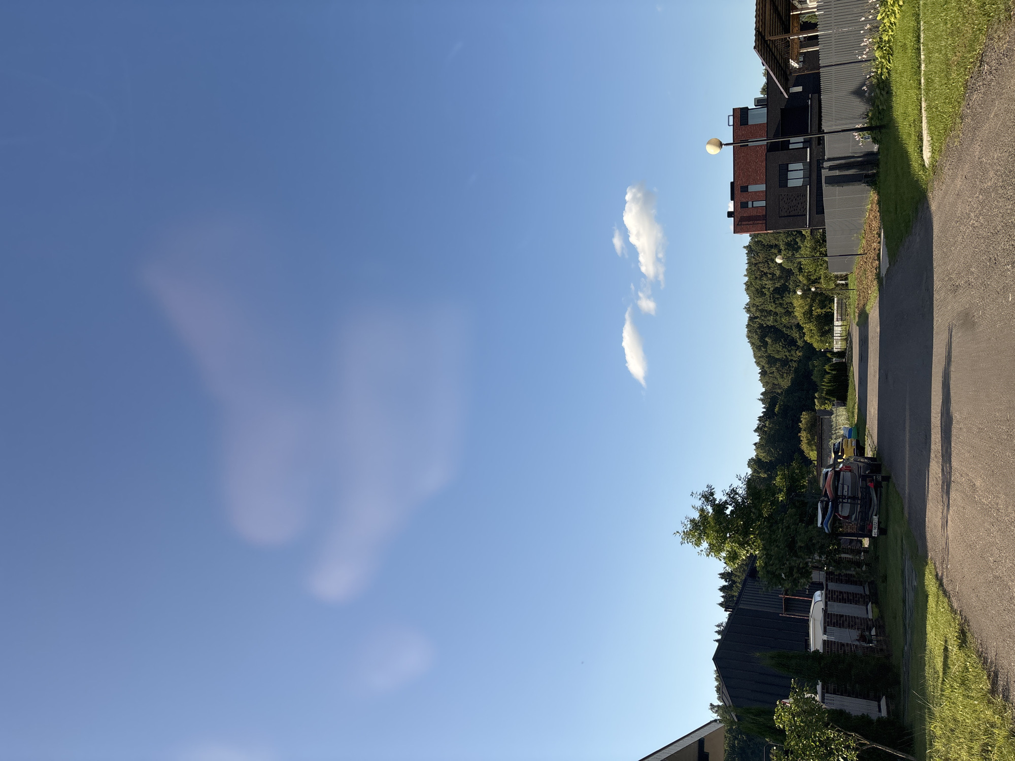 The image depicts a residential street in Zvenigorod, Russia, under a bright blue sky with scattered clouds. The street is paved with asphalt and lined with green grass and trees. On the left side of the street, a modern house with a dark roof and grey fences is visible. Further down, a trailer with kayaks and surfboards is parked next to a yellow car. On the right side, a contemporary house with a dark facade and a red brick upper section stands prominently, with streetlights lining the fence. The background is dominated by a dense forest.