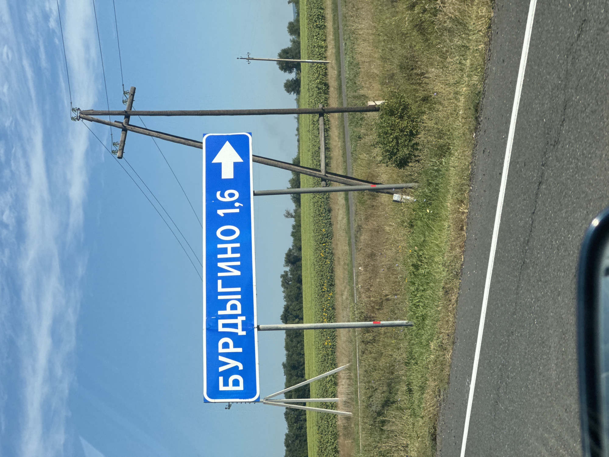 A road sign in Russian that reads "Бурдыгино 1,6" with an arrow pointing right is visible. The sign is blue with white lettering and is mounted on a metal pole. In the background, there is a field of sunflowers, trees, and a clear blue sky with some clouds. There is also a power pole with wires. The foreground shows a portion of an asphalt road with a white line marking. The scene appears to be in a rural area.