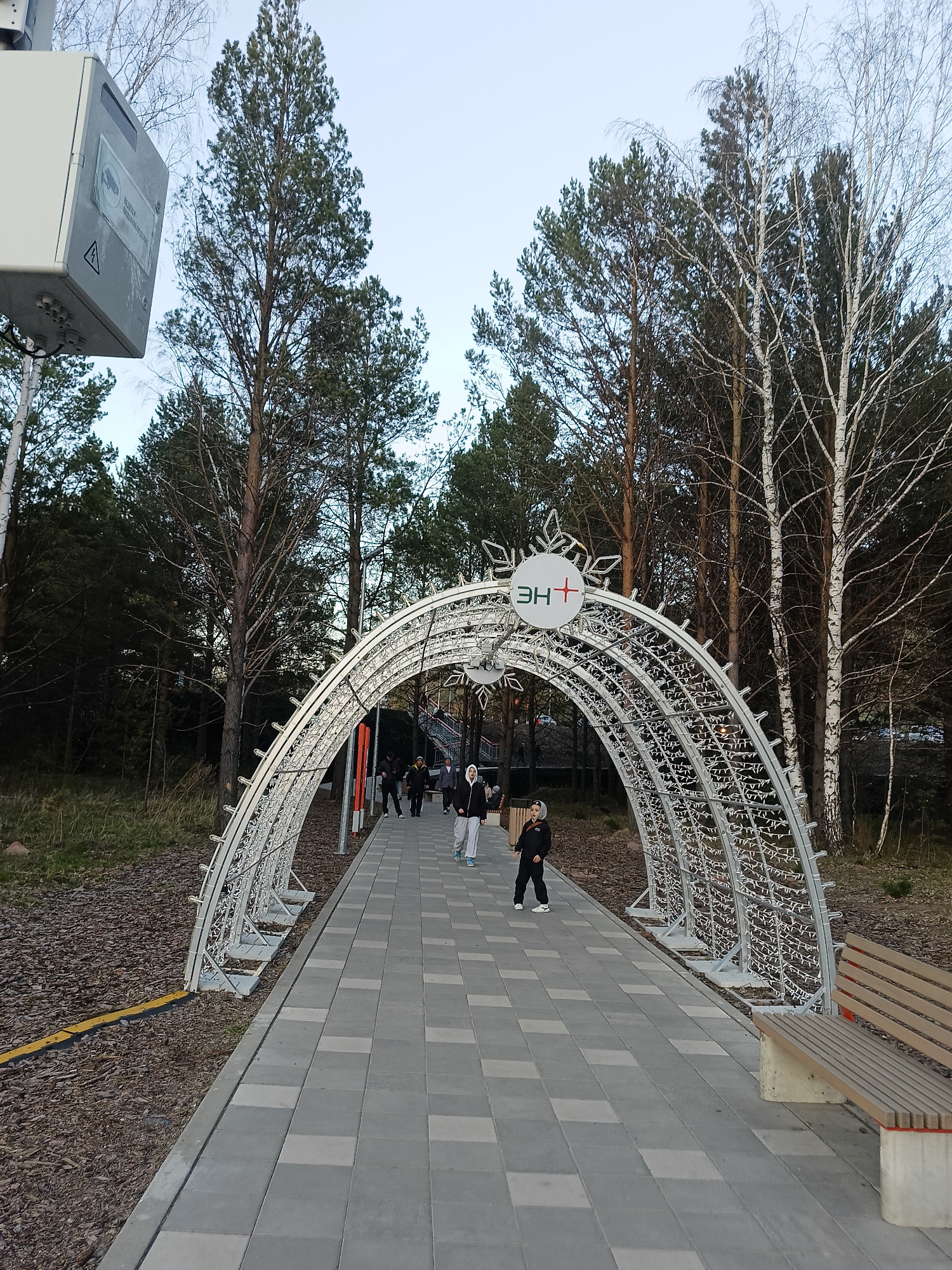 The image depicts a path through a park in Divnogorsk, Russia, likely during the late autumn or winter season. The path is paved with grey rectangular stones and is flanked by tall trees, some of which are deciduous and bare, while others are evergreens. An illuminated arched structure, decorated with small lights and topped with a circular sign bearing the Cyrillic letters "EH" and a red cross, spans the pathway, creating a tunnel effect. Several people are walking along the path, including a child in a dark jacket and light-colored pants. A wooden bench sits on the right side of the path, near the arch. A grey electrical box is visible on the left side of the image. The sky is overcast, suggesting a grey day.