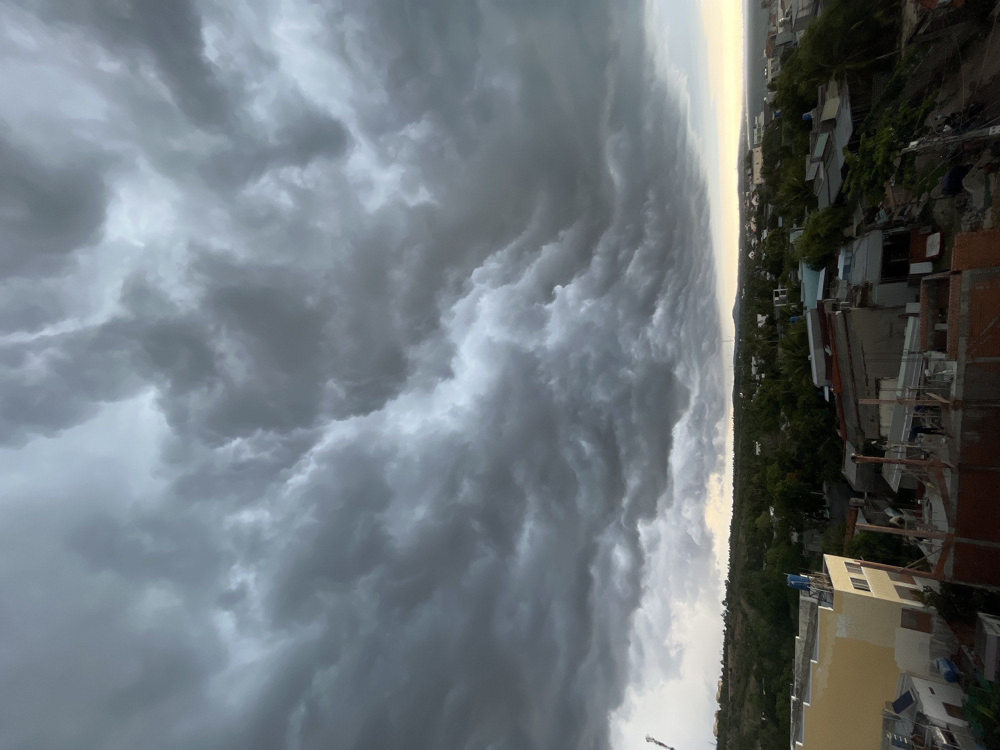 The image depicts a city skyline in Mui Ne, Vietnam, under a dramatic and heavily clouded sky. The clouds are dark and turbulent, with swirling patterns indicating an impending storm or the aftermath of one. Below the sky, a landscape of buildings and lush green foliage is visible. The structures appear to be a mix of residential and possibly commercial buildings, with some under construction, evidenced by exposed brickwork and scaffolding. The architecture is varied, with some buildings featuring light-colored facades, while others have metallic or darker roofs. Trees and other vegetation are abundant, creating a green canopy over parts of the city. In the distance, a hazy horizon suggests the presence of water, likely the sea, given Mui Ne's coastal location. The overall impression is one of a busy, developing urban area set against a powerful natural phenomenon. There are no visible people engaged in specific actions, though some figures might be discernible in the distance or within the buildings, not clearly identifiable. The environment is characterized by a dense, overcast atmosphere and a typical coastal Vietnamese cityscape.