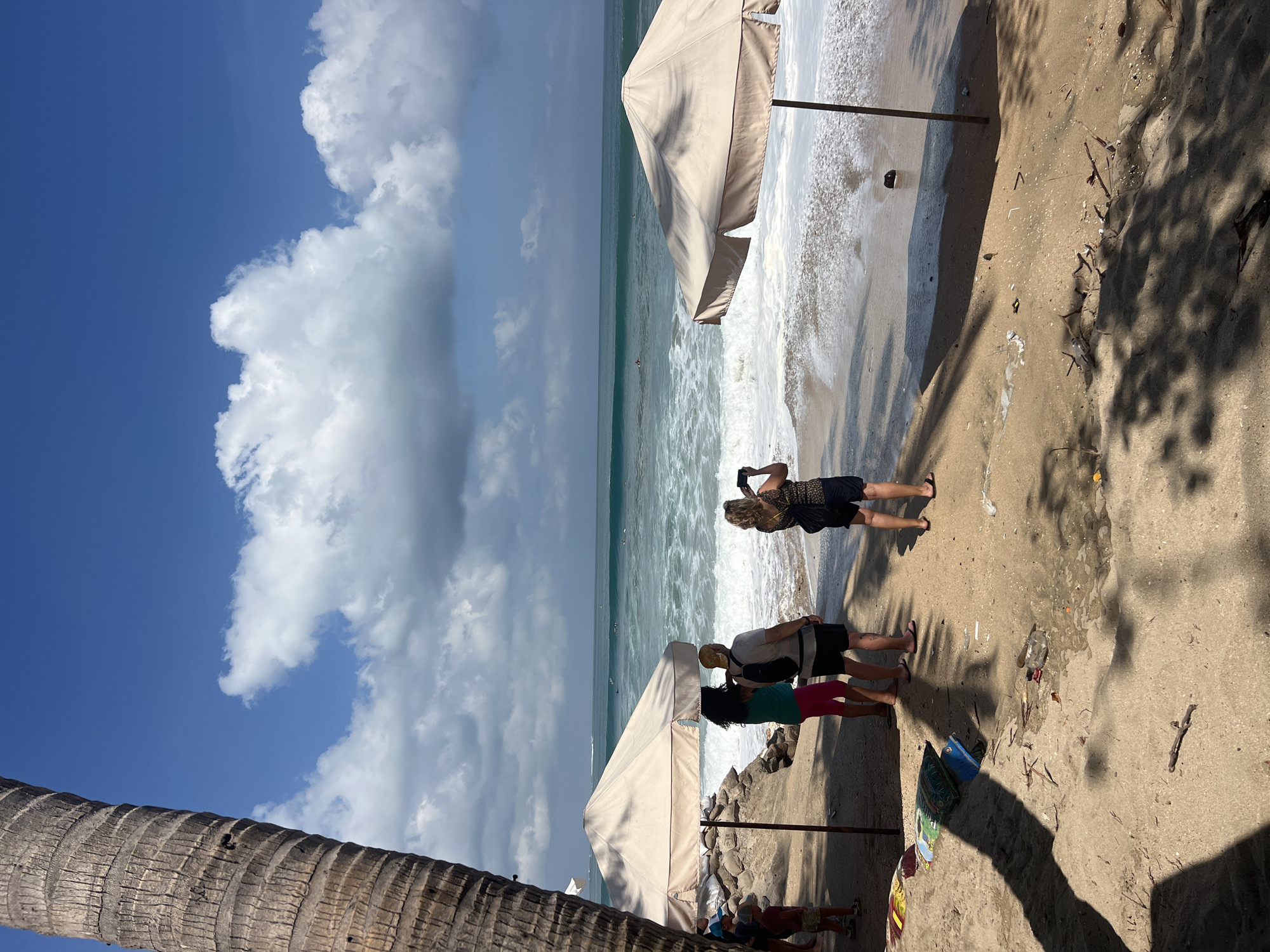 A beach scene in Kuta, Indonesia, is depicted with a focus on the shoreline and the ocean. A prominent palm tree trunk is visible on the left side of the frame, casting a shadow across the sandy beach. In the foreground, several people are standing on the sand, with some appearing to be looking out at the ocean. Two beach umbrellas are set up, providing shade. The ocean exhibits waves and several individuals can be seen in the water, possibly surfing or swimming. The sky is blue with scattered clouds. The sand is littered with small debris.