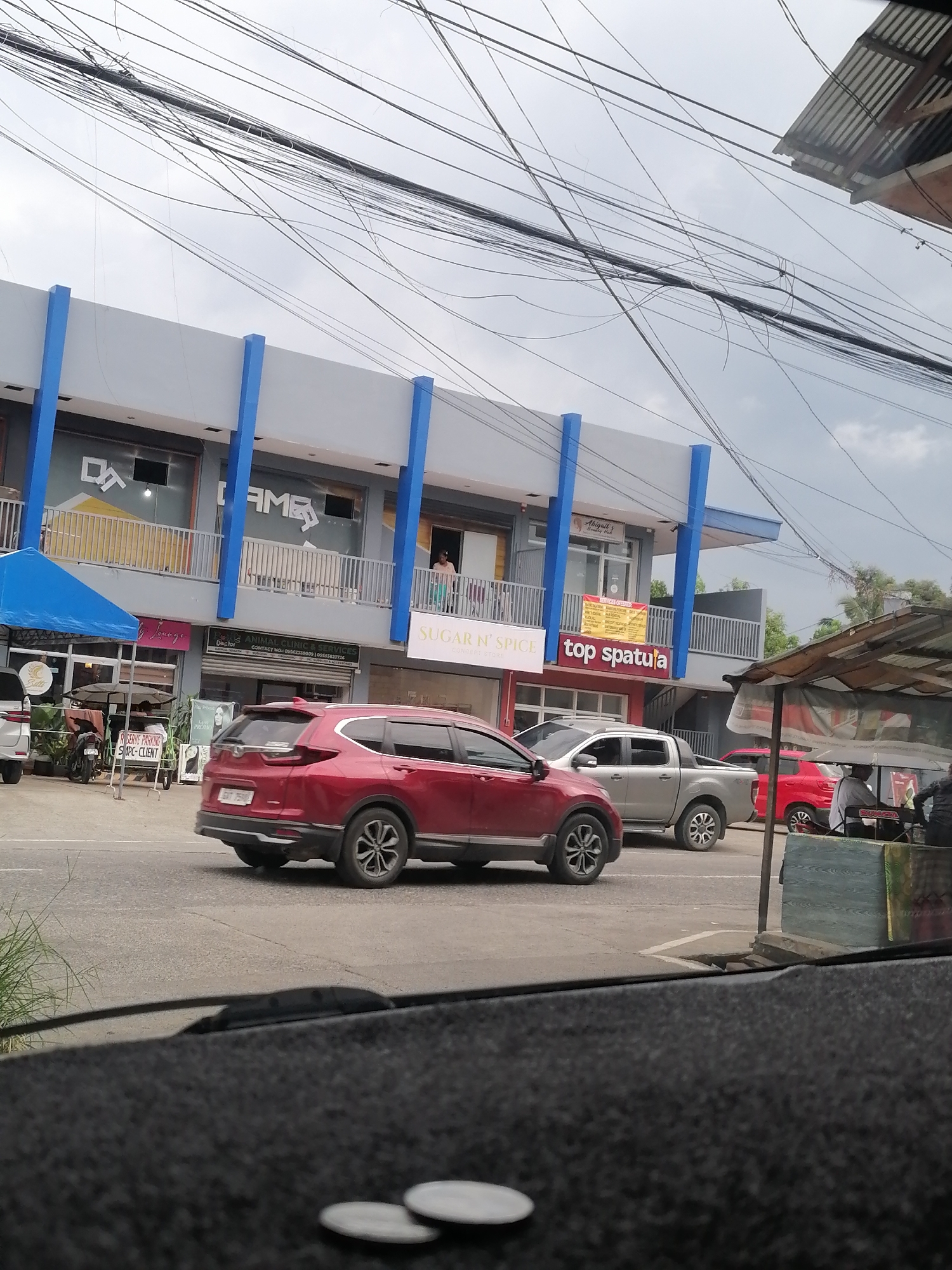The image shows a street scene in Davao City, Philippines. The focal point is a two-story commercial building with a gray facade and blue columns. Several businesses are visible, including "D'AME," "Sugar n' Spice," and "Top Spatula." Signage for "Animal Clinic & Services" is also present. Various vehicles are on the road, including a red SUV (possibly a Honda CR-V), a silver pickup truck, and another red car. A blue canopy is set up in front of one of the businesses. Overhanging wires clutter the sky. Two coins are visible on the dashboard of the vehicle from which the photo was taken.