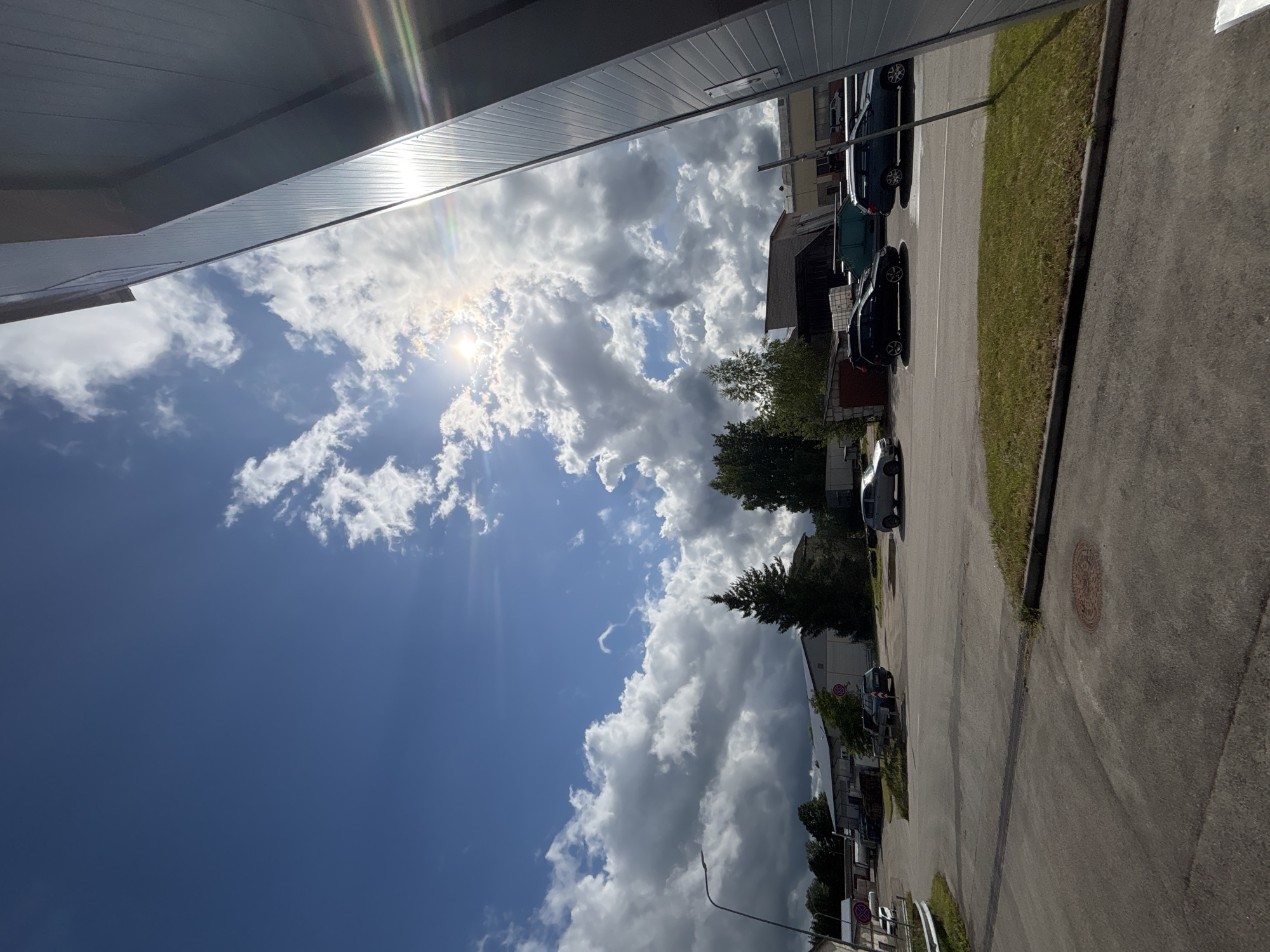 The image depicts an outdoor scene on a sunny day with scattered clouds. A modern industrial building with a corrugated metal facade occupies the right side of the frame, with the sun creating lens flare and reflections on its surface. The foreground is an asphalt road and a patch of green grass. In the background, several cars are parked. Beyond the parking area, there are industrial buildings, trees, and a prominent evergreen tree. The sky is a vibrant blue with bright white and dark grey clouds, indicating dynamic weather. The location appears to be in an industrial area, possibly on the outskirts of Sauleskalns, Latvia, given the architectural style and the presence of industrial facilities. There are no people visible in the image.