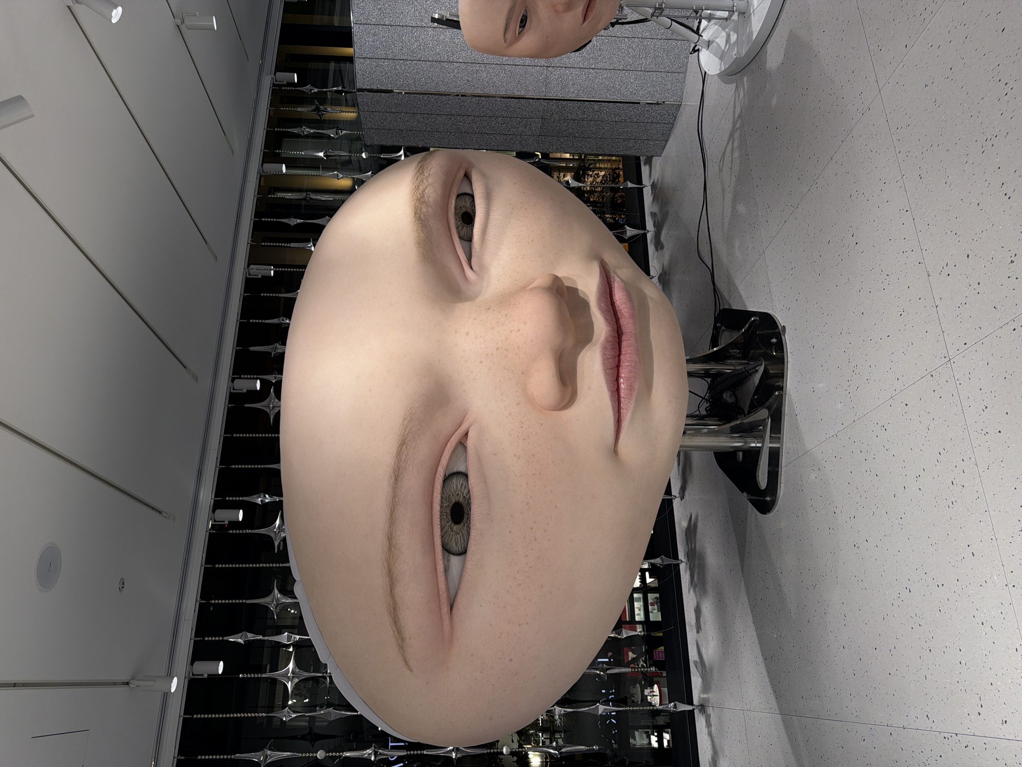 A close-up, eye-level shot showcases a large, hyperrealistic mannequin head with a round, smooth forehead and visible freckles. The mannequin's left eye, facing the camera, is a detailed iris of green and brown hues, with a slightly downturned gaze. The right eye is partially visible and also appears to be looking slightly down. The lips are a soft pink, with a subtle, closed-mouth expression. The mannequin is mounted on a metal stand, with another smaller mannequin head partially visible in the background to the right. The environment appears to be an indoor exhibition space, with a speckled grey floor and a backdrop of decorative metallic hanging elements and a wall panel. The lighting is overhead, with spotlights visible. The scene is located in the retail and exhibition district of Chuo, Japan.