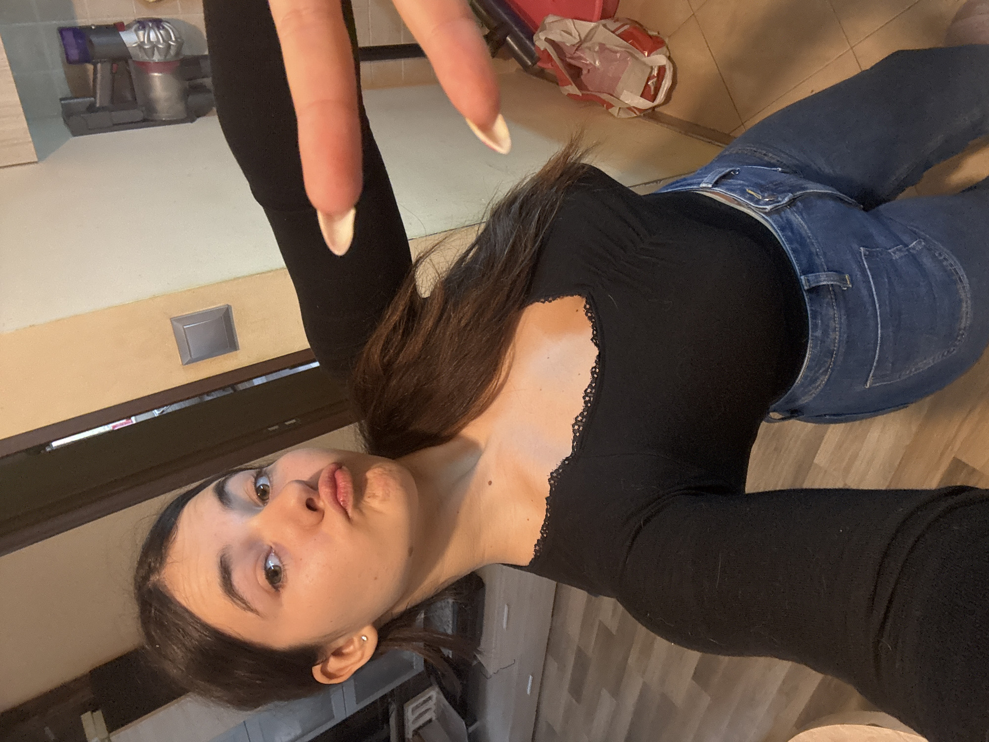 A young woman is depicted in a casual indoor setting, appearing to be lying down while taking or posing for a photo. Her head is tilted, and she looks directly at the camera with a playful "fish face" or pout, her lips pursed. She has dark brown hair, parted to the side, and a small, light-colored stud earring in her left ear. She is wearing a fitted black long-sleeved top with a delicate lace trim along the neckline, paired with blue jeans. Her fingernails are manicured with a light polish.
The background suggests a domestic environment, likely a kitchen or living area in Plovdiv, Bulgaria. Visible behind her head and shoulders is a light-colored wall with a darker trim, featuring a small gray rectangular object, possibly an electrical outlet or switch. Further in the background, a Dyson handheld vacuum cleaner (purple and silver) is charging or stored on what looks like a counter or shelf. Wooden floorboards are visible in various parts of the image, and a red and white plastic bag, possibly containing household items or refuse, can be seen on the floor. The overall impression is one of a relaxed, informal moment captured indoors. No explicit text is visible in the image.