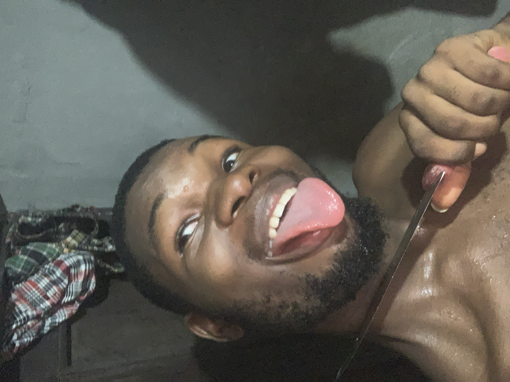 A dark-skinned man with a beard is lying on his back, facing upwards and to the right of the frame. He has his mouth open, tongue extended. He appears sweaty. A silver knife is positioned near his neck. A hand grips a part of his body near the knife. A plaid cloth is in the background. The location and the city of Onitsha, Nigeria cannot be determined from the image.