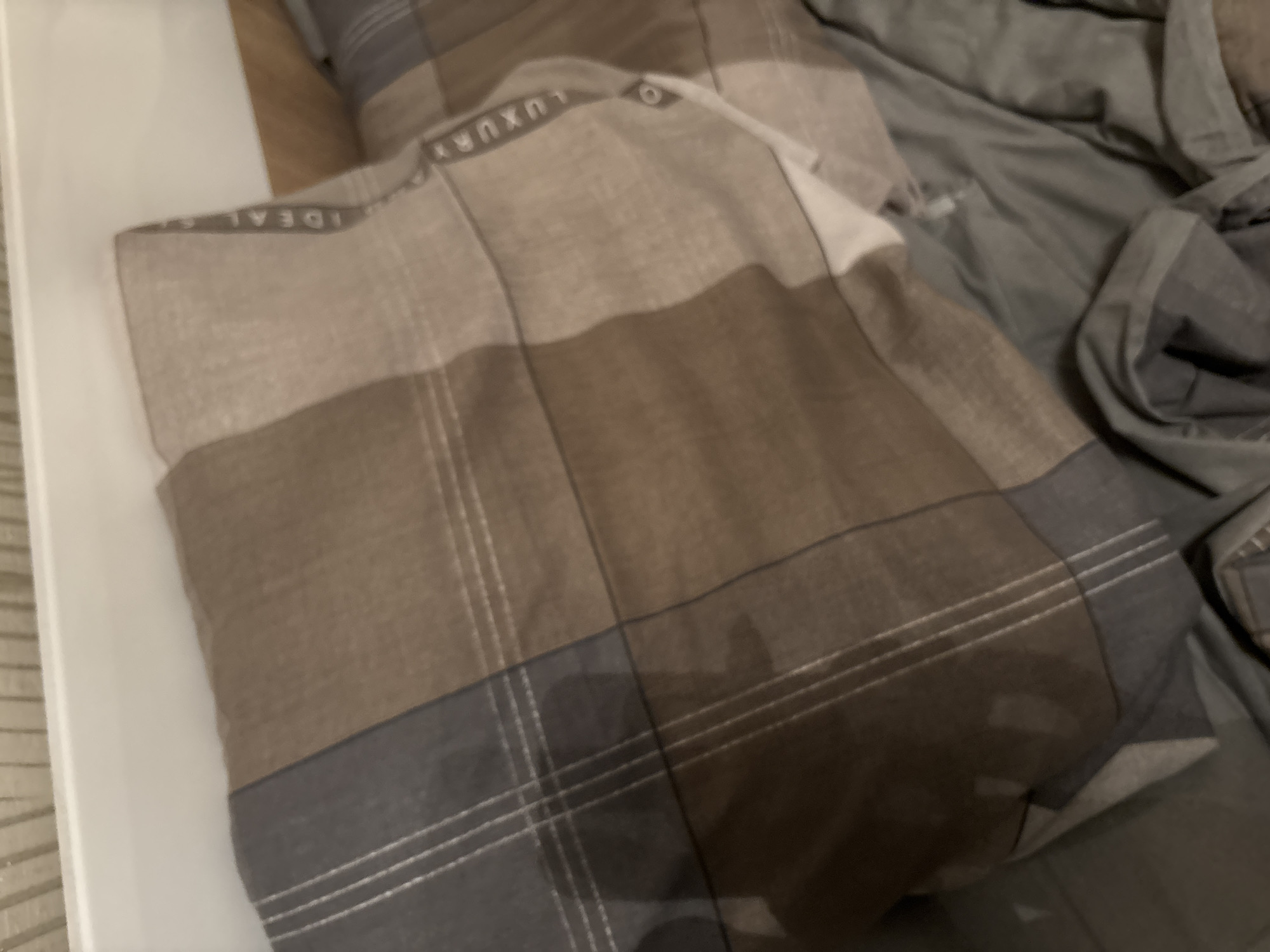 This image, taken in Chernogorsk, Russia, displays an indoor scene focused on bedding. A rectangular pillow is centrally positioned, covered with a plaid pillowcase featuring a pattern of alternating brown, beige, and dark grey-blue squares. Thin white lines form a grid across the plaid design. Vertically oriented text, "IDEAL" and "LUXURY," is visible on one of the dark grey-blue bands of the pillowcase. To the right and below the pillow, crumpled fabric in a solid dark grey-green hue is visible, consistent with a duvet or sheet. In the upper left background, a textured light-colored surface, possibly a headboard or wall, is present, topped by a smooth white surface. No individuals or active subjects are depicted.
