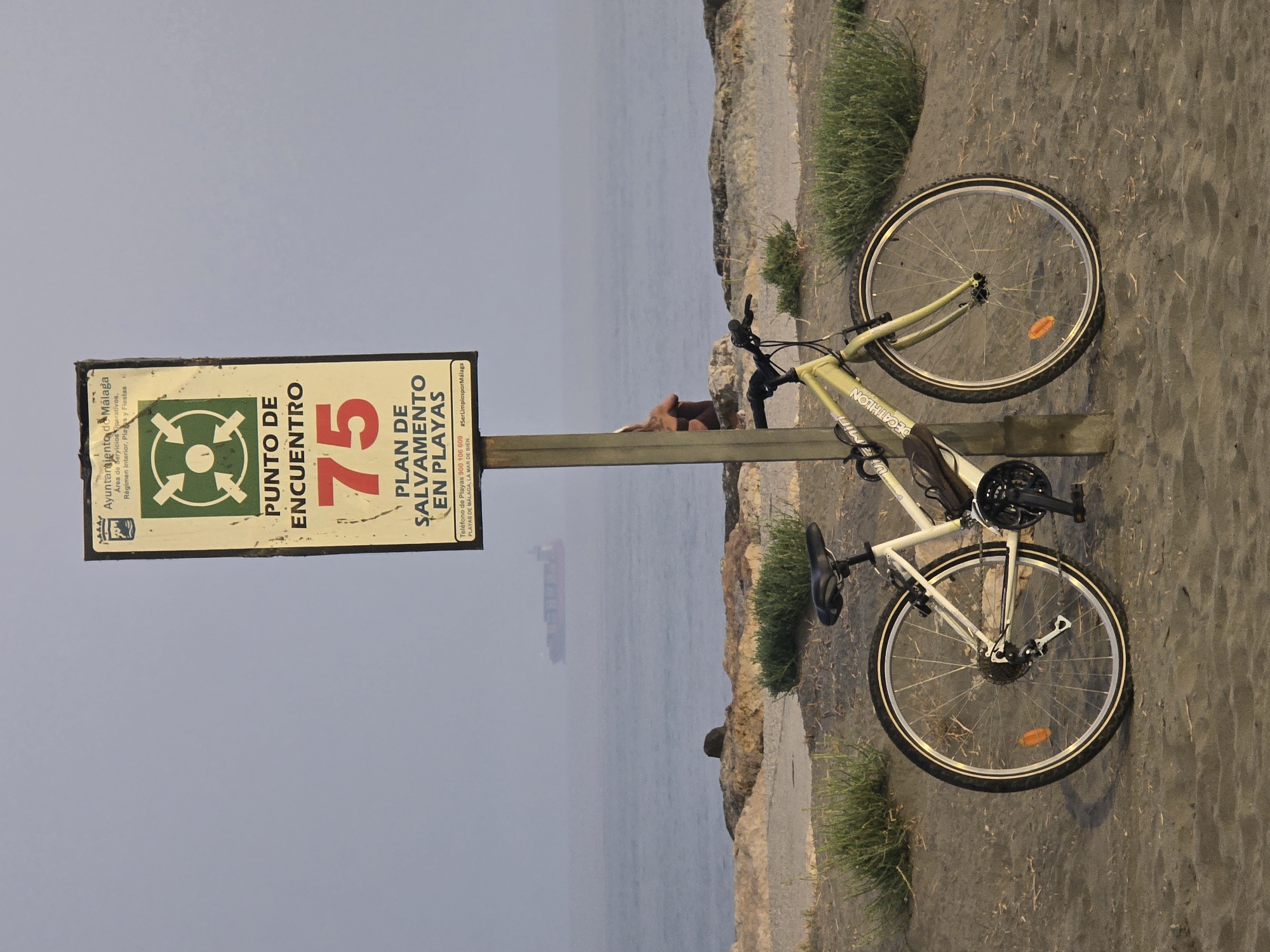 An outdoor, eye-level, medium shot captures a bicycle parked on a sandy beach in Málaga, Spain, next to a pole with a sign. The sign, mounted on the pole, features a green symbol of four arrows pointing inward towards a central circle, with text below indicating "PUNTO DE ENCUENTRO 75" (Meeting Point 75) and "PLAN DE SALVAMENTO EN PLAYAS" (Beach Rescue Plan). The bicycle, a light yellow and white model, is positioned in front of the sign, with its front wheel slightly turned to the left. A person is visible in the background, sitting on rocks near the water. The sea stretches out behind the beach, with a large cargo ship visible in the hazy distance. Green vegetation grows along the edge of the sand and rocks. The lighting suggests it is either early morning or late evening, with a muted, overcast sky.