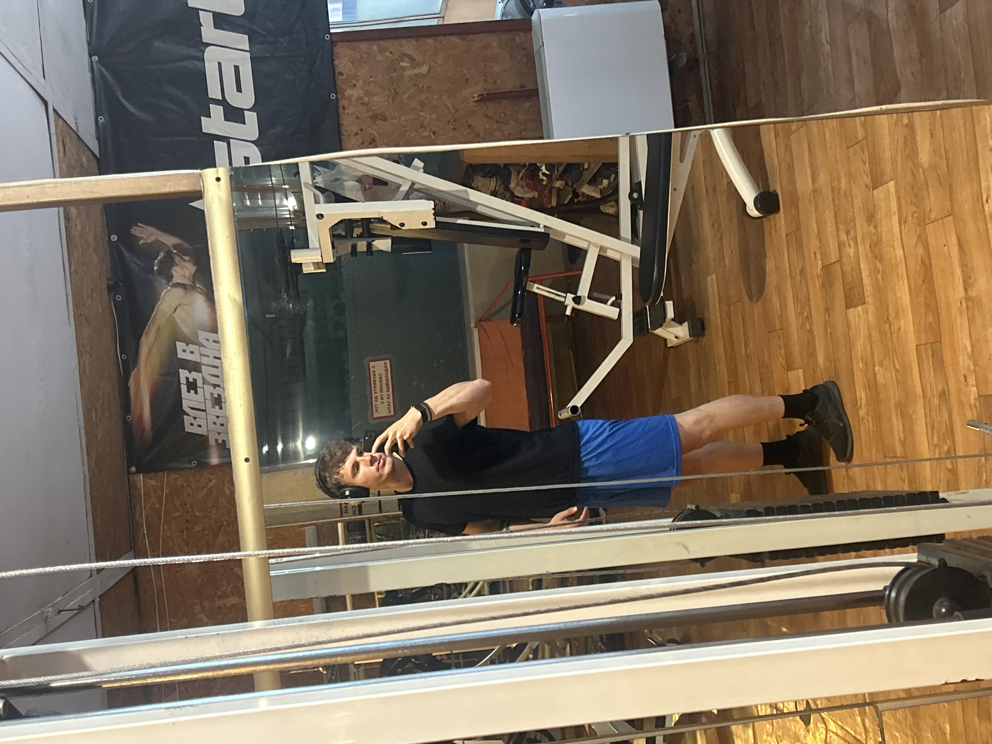 The image is taken indoors, presumably in a gym, evidenced by the presence of various exercise machines. A man is visible in the foreground, seemingly holding a phone to his ear, possibly on a call. He is wearing a black t-shirt, blue shorts, black socks, and black shoes. A banner with the word "Start" is visible in the background. The location is in the city of Elhovo, Bulgaria, as indicated by the Cyrillic text on the banner. The gym appears to have wooden floors and walls.