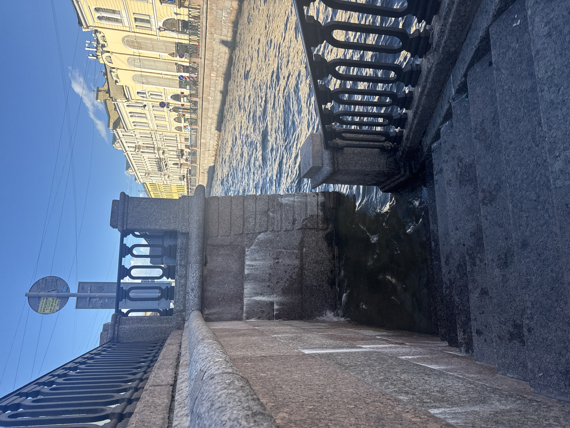 This is a medium-angle, full-shot photograph of a canal in Saint Petersburg, Russia, under a clear blue sky. In the foreground, a stone embankment curves to the left, with a set of dark grey stone stairs descending into the water on the right. A black metal railing with ornate scrollwork runs along the top of the embankment and the stairs.
The canal itself is a deep, dark blue, with choppy waves reflecting the sunlight. On the opposite bank, a row of yellow buildings with classical architecture lines the canal. These buildings feature many windows and decorative elements. A few people are visible walking along the embankment across the canal, and some small boats or barges are moored near the buildings. Overhead, power lines crisscross the sky.
A circular sign with Russian text is mounted on a pole near the top of the embankment on the left. The overall impression is one of urban architecture and waterway, bathed in bright daylight.
