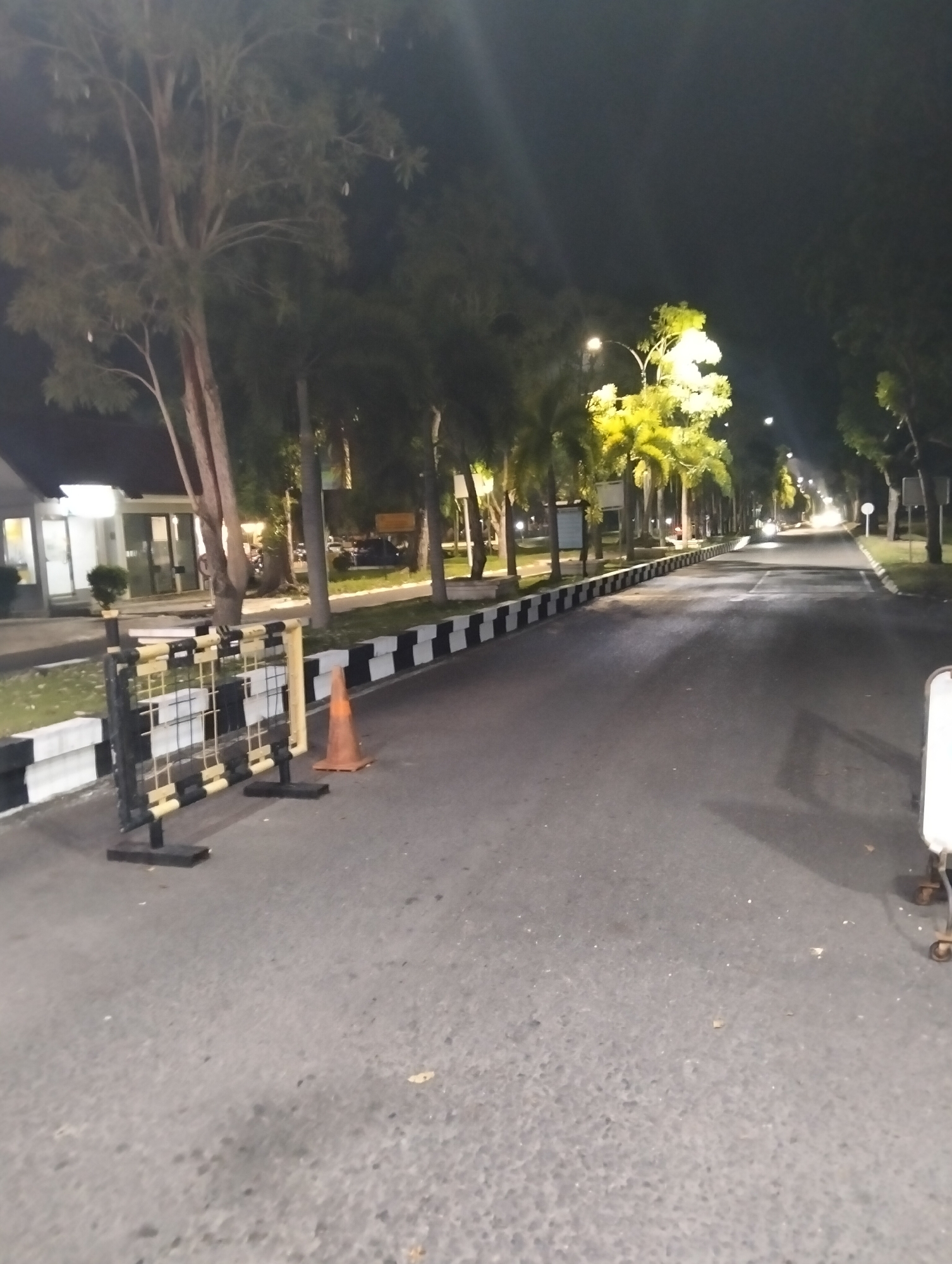 The image captures a nighttime scene on an asphalt road bordered by trees and a white and black striped barrier. In the foreground, a black and yellow striped barricade stands on the left, accompanied by an orange traffic cone. Further down the road, palm trees and other foliage line the sides, illuminated by streetlights. The road stretches into the distance, curving slightly to the right, with more streetlights visible along its path. On the left side of the road, a building with windows and lights can be seen. The overall setting appears to be an outdoor area, possibly a campus or a park, in Tambontunong, Indonesia, as indicated by the location context. The time of day is clearly nighttime, indicated by the dark sky and artificial lighting. No people are visible in the frame. The image conveys a sense of quietness and emptiness due to the absence of activity and people. No visible text is present in the image.