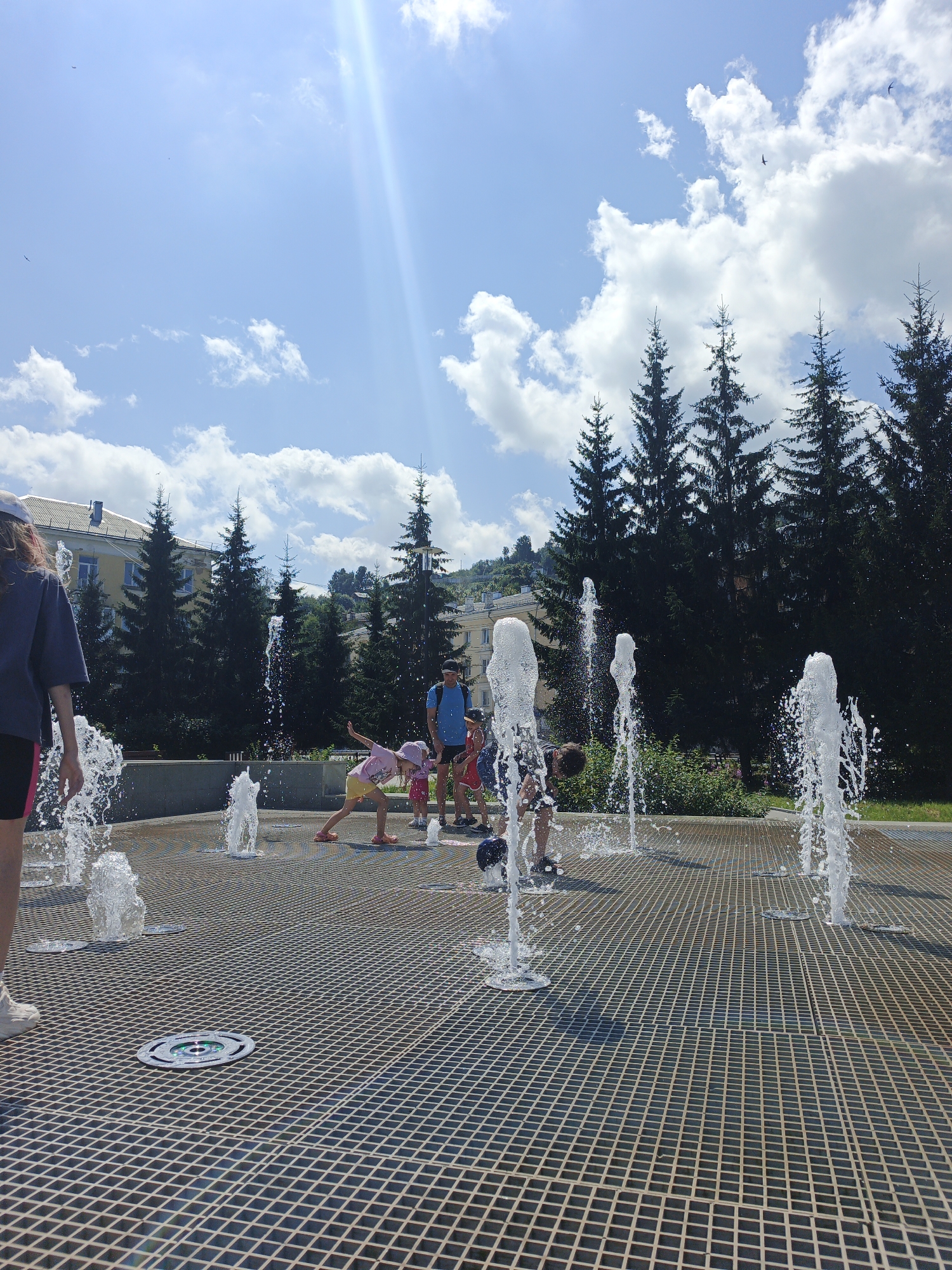 In Zlatoust, Russia, on a sunny day with a bright blue sky dotted with clouds, a group of people, predominantly children, are enjoying a water fountain display in an urban park. The fountain features multiple jets of water spraying from a metal grate surface. Several children are actively engaged with the water, some running through it, others bending down, and one is captured in mid-air, appearing to jump or play. A few adults are also present, observing the children. The background reveals a line of evergreen trees, urban buildings, and a hillside. The overall scene depicts a recreational activity in a public space.