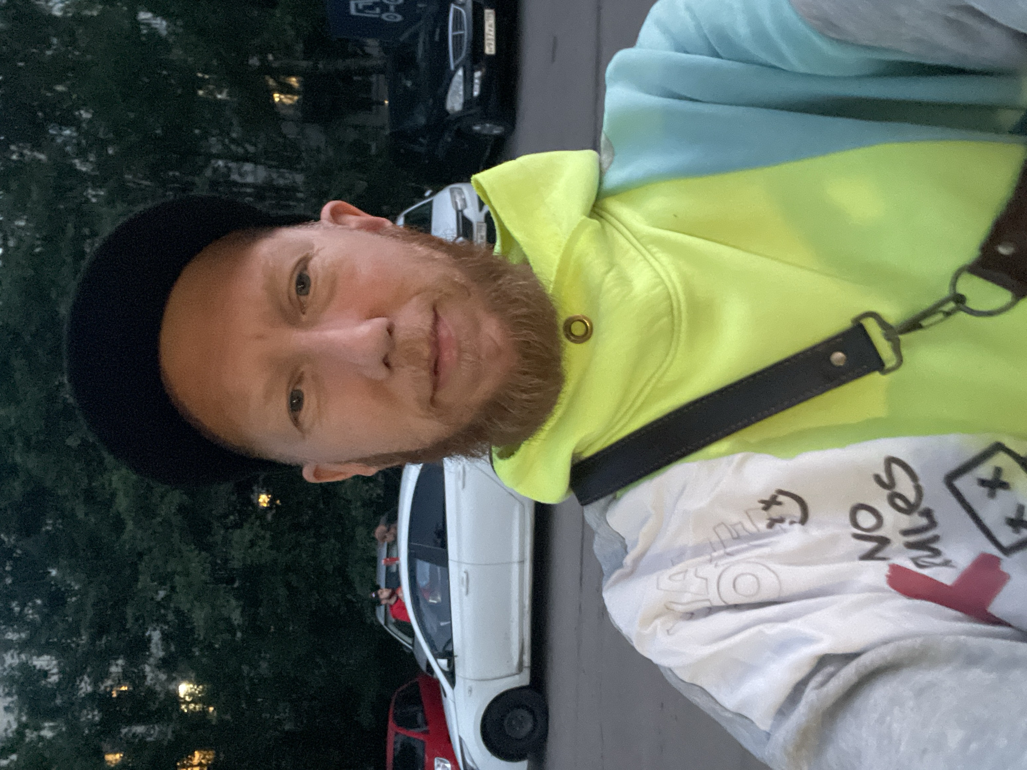 The image is a selfie taken by a man with a beard and a black baseball cap. He is wearing a brightly colored hooded sweatshirt. In the background, there are several cars parked on a paved surface. The environment appears to be an urban setting with trees and buildings visible in the distance, suggesting a park or residential area within the city of Saint Petersburg, Russia. The lighting suggests it is late evening or early morning.