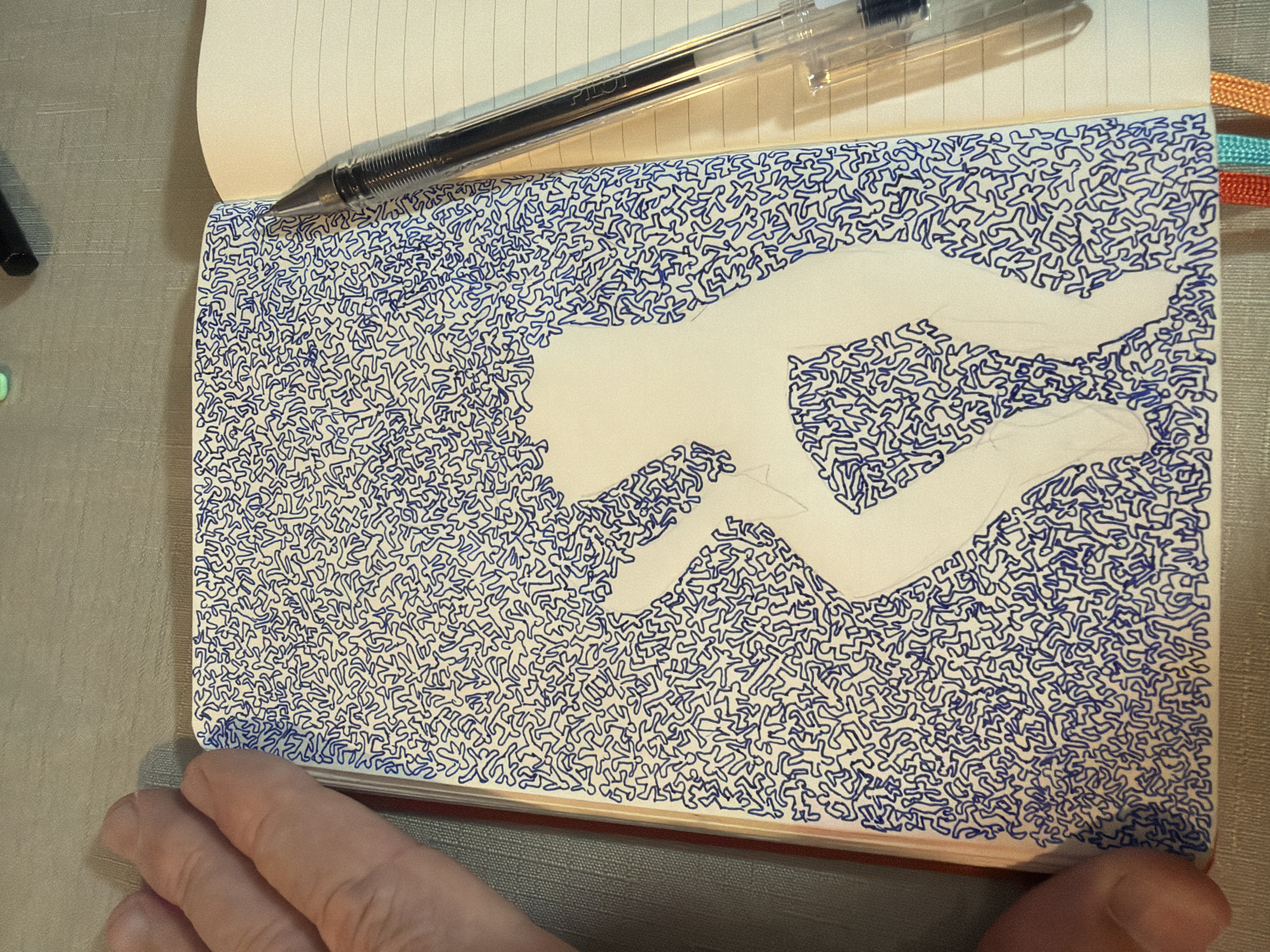 A hand and a pen are visible in the foreground of an open notebook. The notebook's left page is filled with a pattern of small, blue, stick-figure-like shapes. In the center of this page, a silhouette of a person is drawn in white, with faint pencil lines visible inside the silhouette. The right page of the notebook is lined. A black pen rests on the right page, near the spine of the notebook. The background consists of a textured, light-colored surface. The scene appears to be indoors, on a table. The city of Even Yehuda, Israel is not depicted or suggested by any elements in the image.