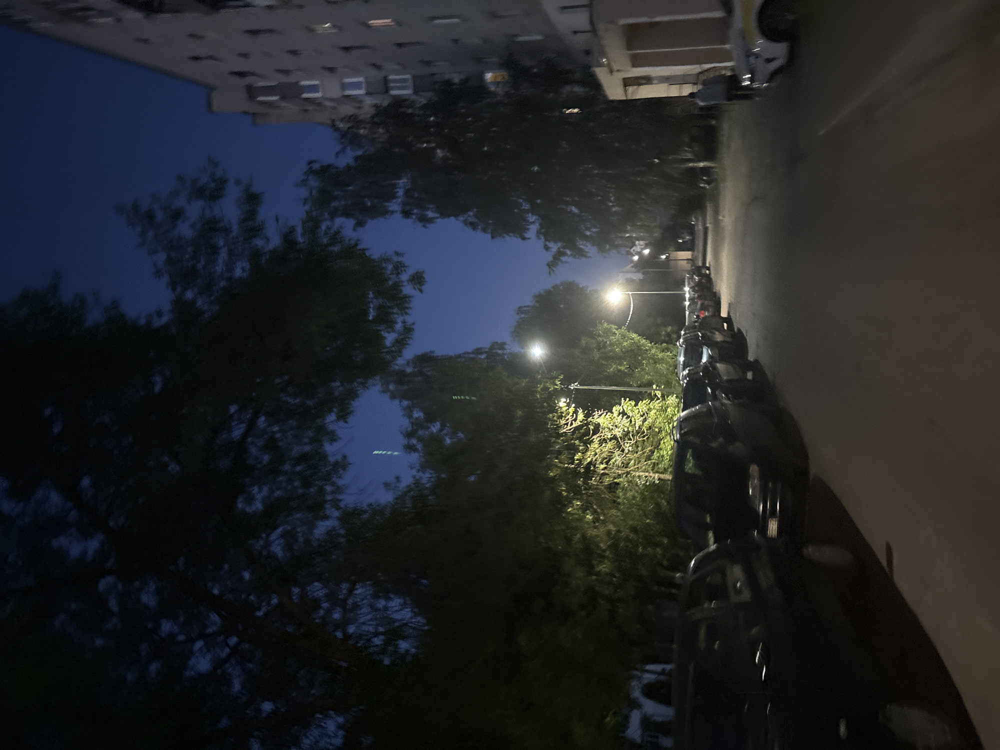 The image depicts a residential street scene at dusk or night in Vitsebsk, Belarus. On the right side of the street is a multi-story apartment building with visible lit windows, indicating inhabitants. The building's facade is plain and grey. On the left side of the street, parked cars are visible, mostly dark-colored and appearing to be sedans or SUVs. Trees with dense foliage line the street, with some branches obscuring the view of the sky, which is a deep blue. Streetlights are present, casting pools of light on the street and illuminating the trees. One streetlight is particularly bright and appears to be near the center of the image. In the distance, the street recedes and appears to have more parked cars and trees. A person is walking away from the camera on the sidewalk in front of the apartment building. Another white car with yellow markings, possibly a service vehicle, is parked further down the street. The overall atmosphere is quiet and typical of a residential area after dark.