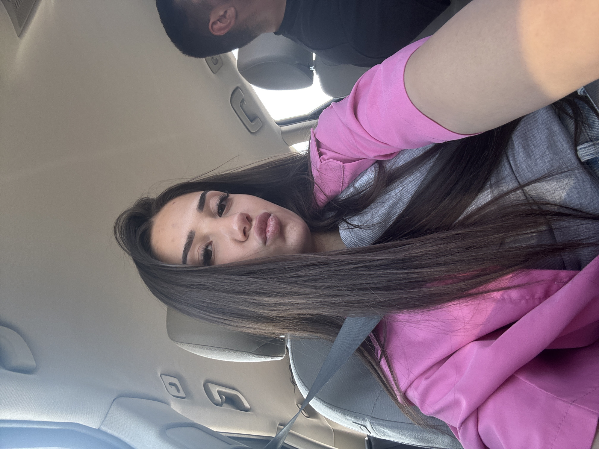 The image is a selfie taken inside a car, oriented on its side. The primary subject is a young woman, positioned in the lower left half of the frame, looking directly into the camera. She has long, straight dark brown hair that cascades over her left shoulder and chest. Her face shows a slight pout, with prominent eyelashes and defined eyebrows. She is wearing a bright pink long-sleeved top or jacket over a gray garment, possibly a hoodie, which is visible around her neck and arm. A dark gray seatbelt is fastened across her chest and lap.
In the upper right background, partially cropped, the head of a man is visible, wearing a dark shirt. The setting is the interior of a car, with light-colored ceiling and headrests visible. Bright natural light streams in from a window or sunroof located towards the top of the frame, indicating it is daytime. No text is visible in the image. The overall impression is a casual, in-car moment captured as a selfie.