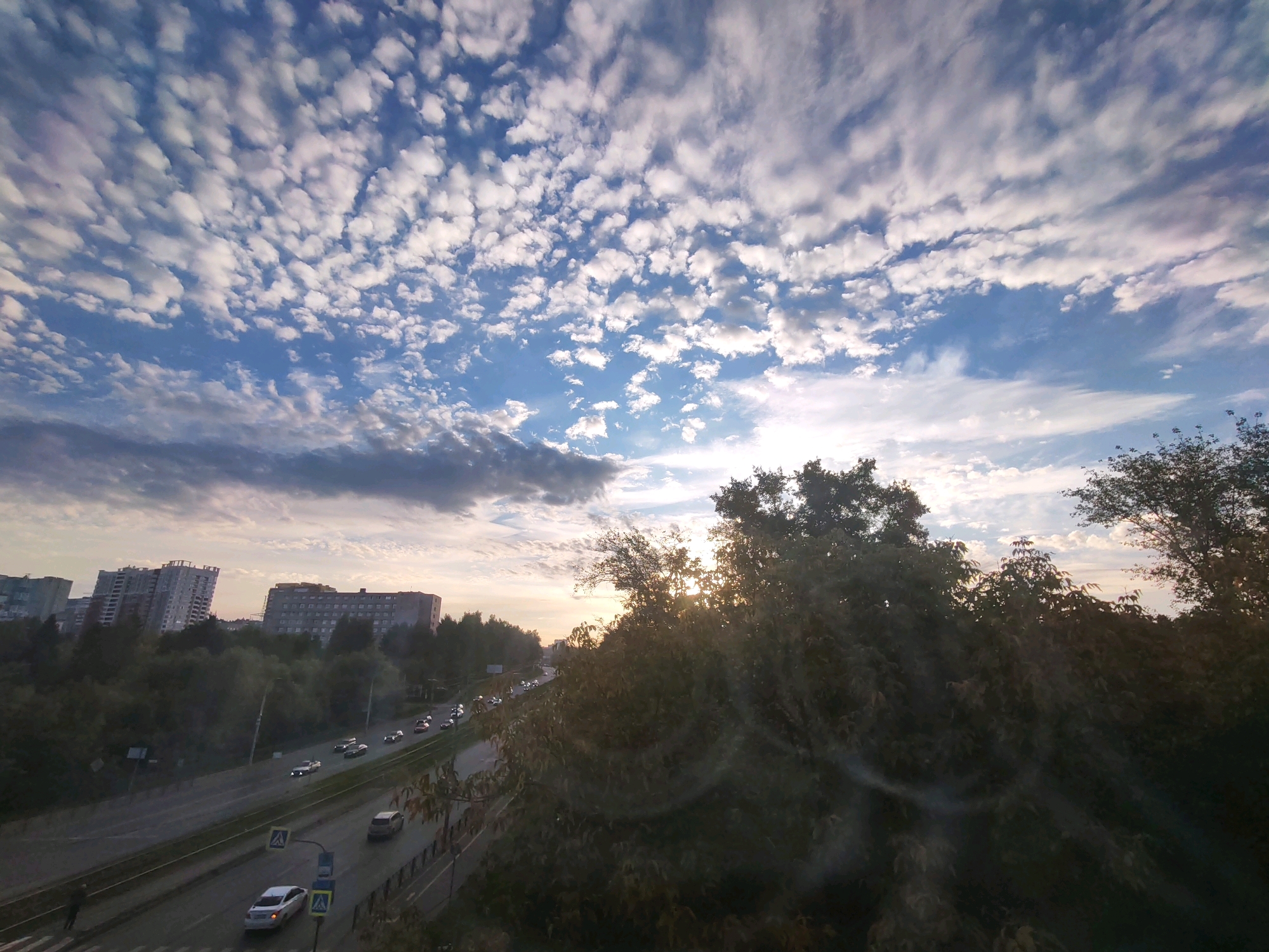 The image captures a view of a multi-lane highway in Izhevsk, Russia, during what appears to be either sunrise or sunset, given the lighting. The sky is filled with scattered, fluffy white clouds against a blue backdrop. In the foreground, dense foliage obscures part of the view, with sunlight filtering through the leaves. Beyond the foliage, several multi-story apartment buildings are visible in the middle ground, indicating an urban environment. The highway itself is populated with a moderate amount of vehicle traffic. A person can be seen walking on a pedestrian crossing on the left side of the road. The overall scene depicts a typical urban landscape with elements of nature and human activity.
